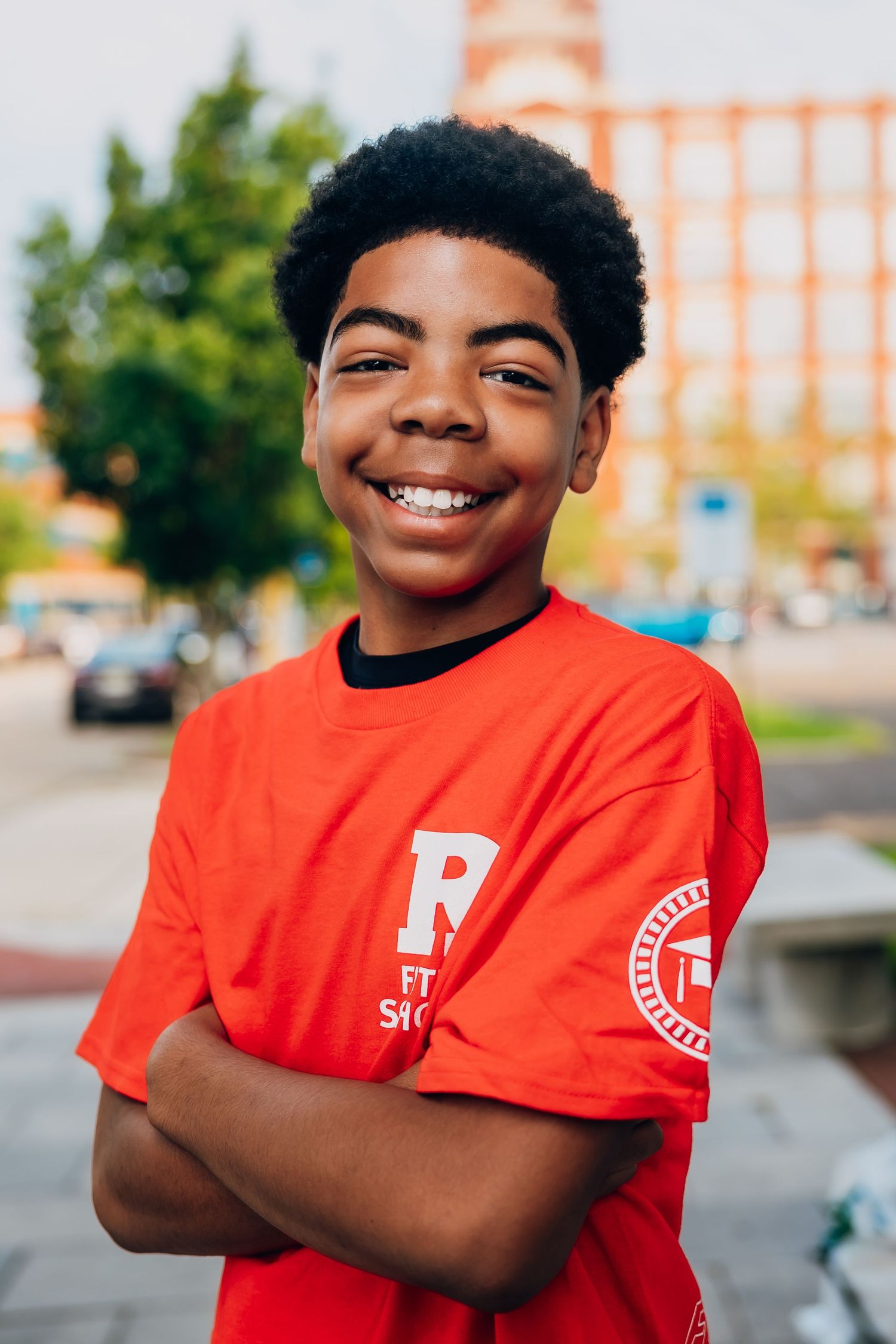 Smiling young boy in red R t-shirt with arms crossed, standing outdoors on a city street.