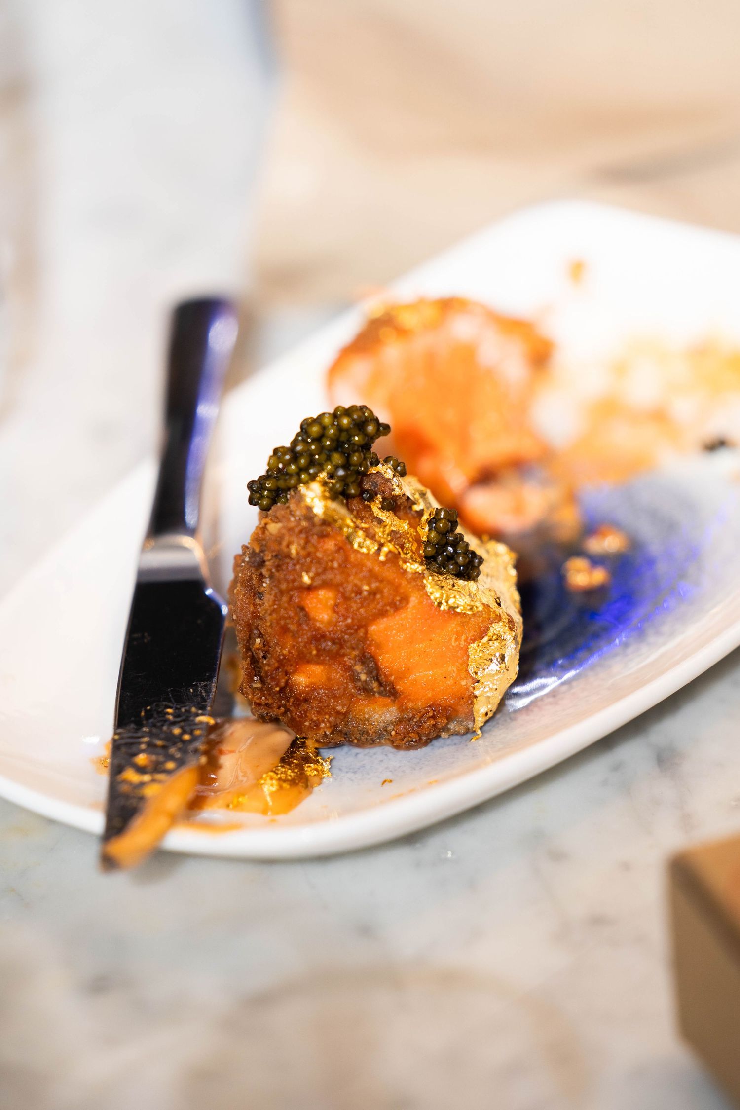 Elegant fried croquette topped with black caviar and edible gold leaf served on a white plate with a knife.