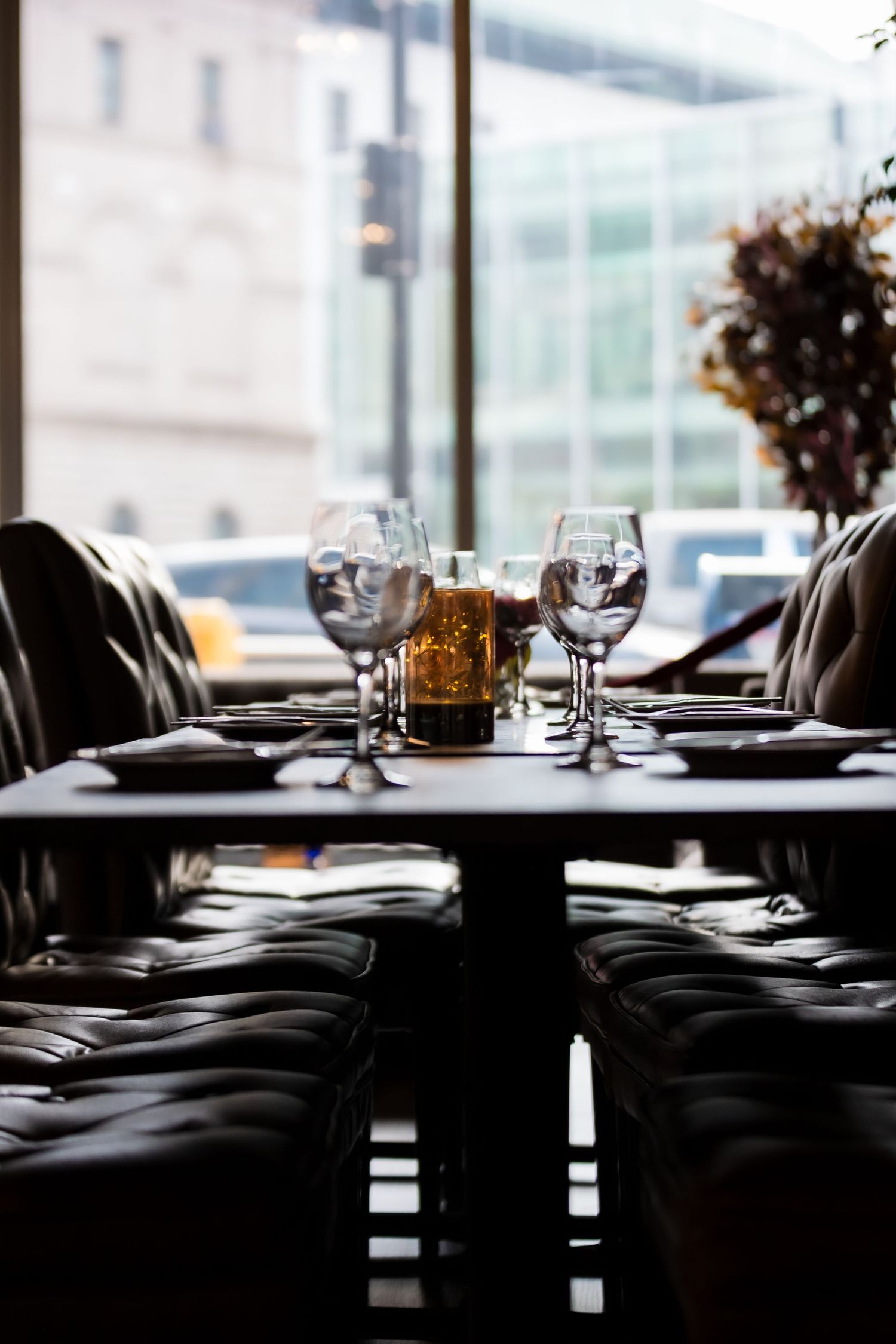 Elegant restaurant table set with wine glasses and candle, leather booth seating, city view through large windows.
