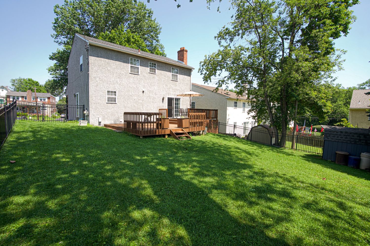 Spacious backyard with green lawn, large wooden deck, mature trees, and privacy fencing on a sunny day.
