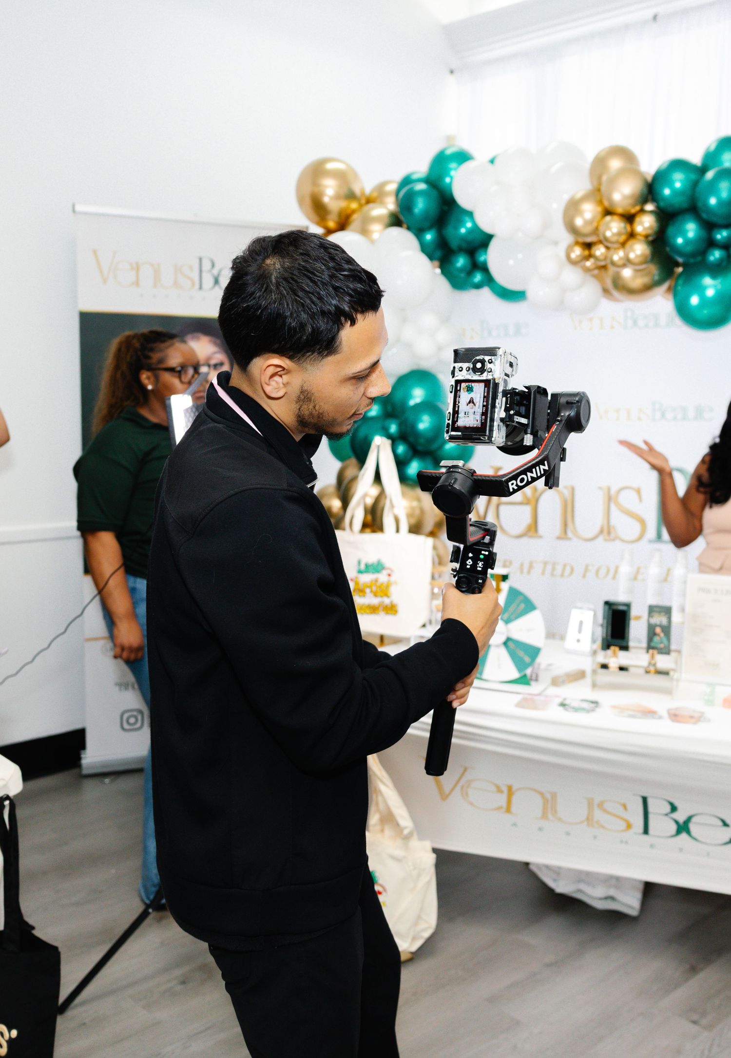 Man filming a VenusBeauty product display table with a camera gimbal at a green and gold balloon event.