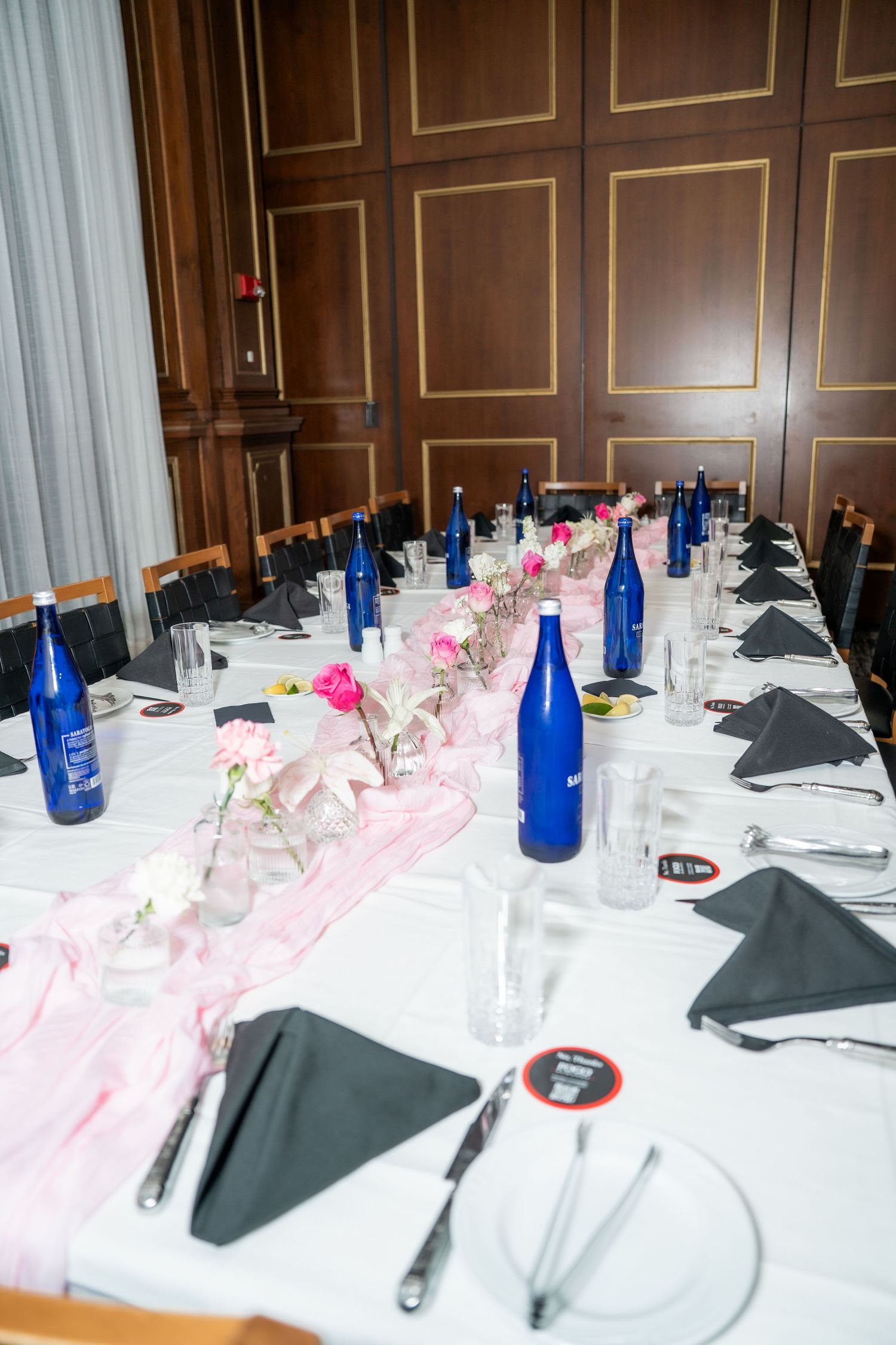 Elegant banquet table set with white linens, pink runner, blue Saratoga water bottles, and floral centerpieces in a wood-paneled room.