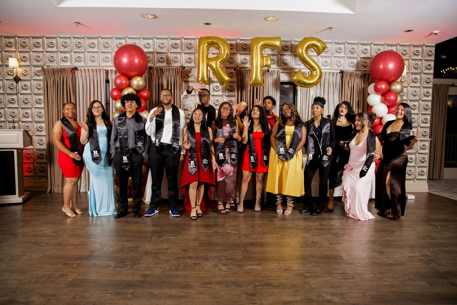 Group of elegantly dressed students posing at RFS school prom with gold letter balloons and red decorations.