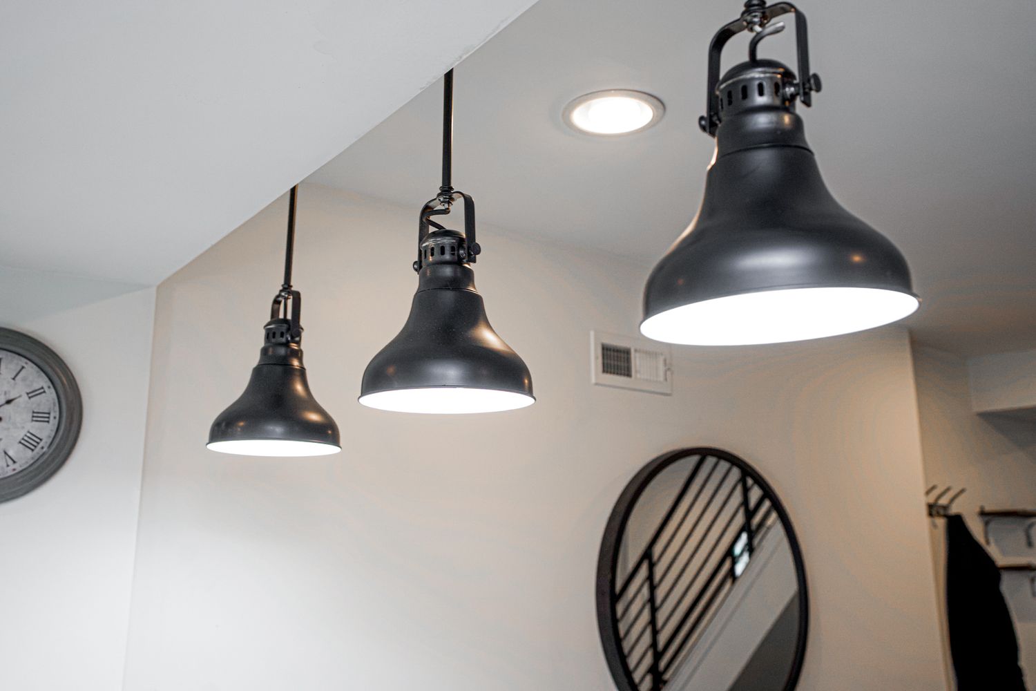 Three black industrial pendant lights hang from a white ceiling near a round mirror and wall clock.