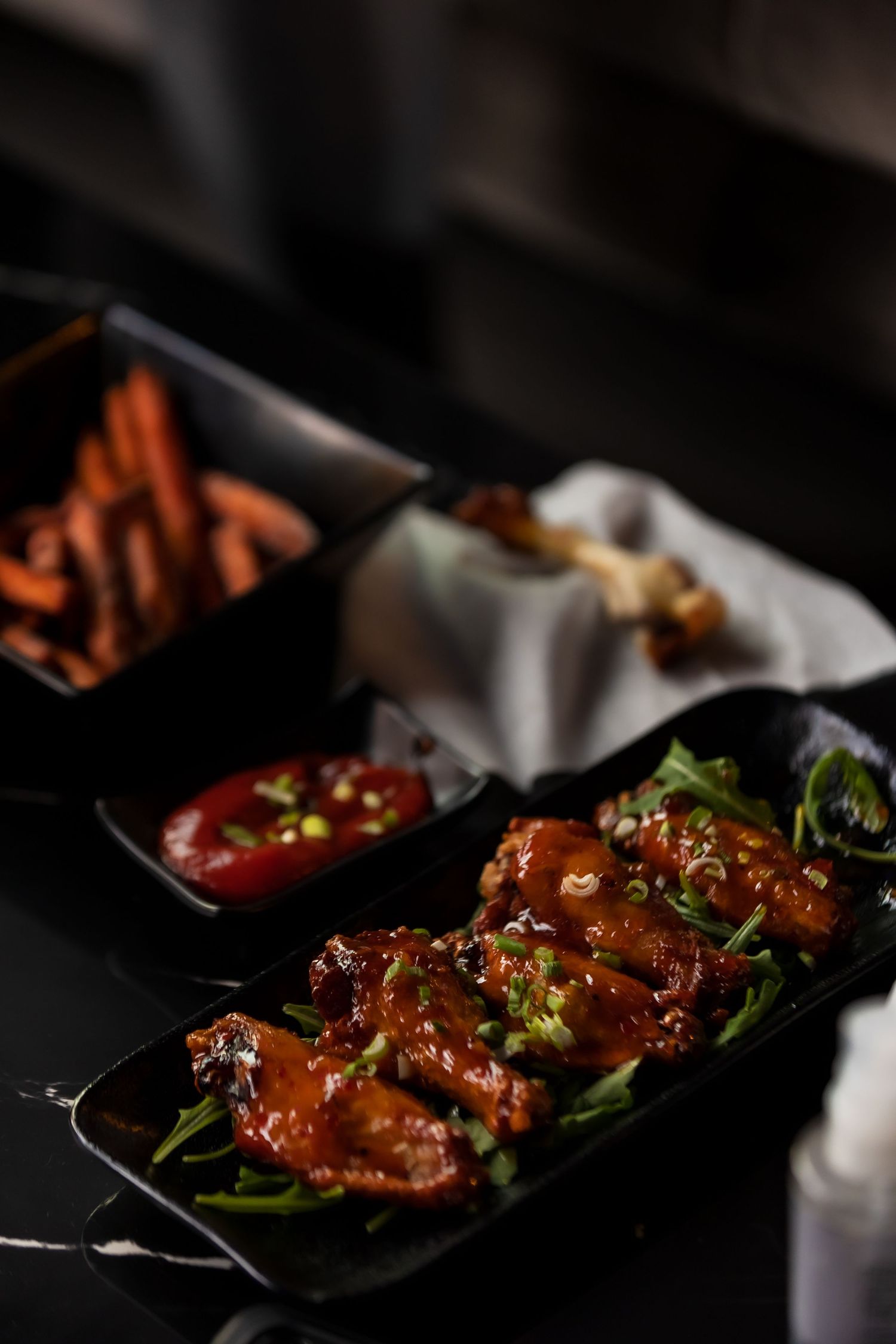 Glazed chicken wings with sesame seeds and green onions served on a black plate with sweet potato fries.