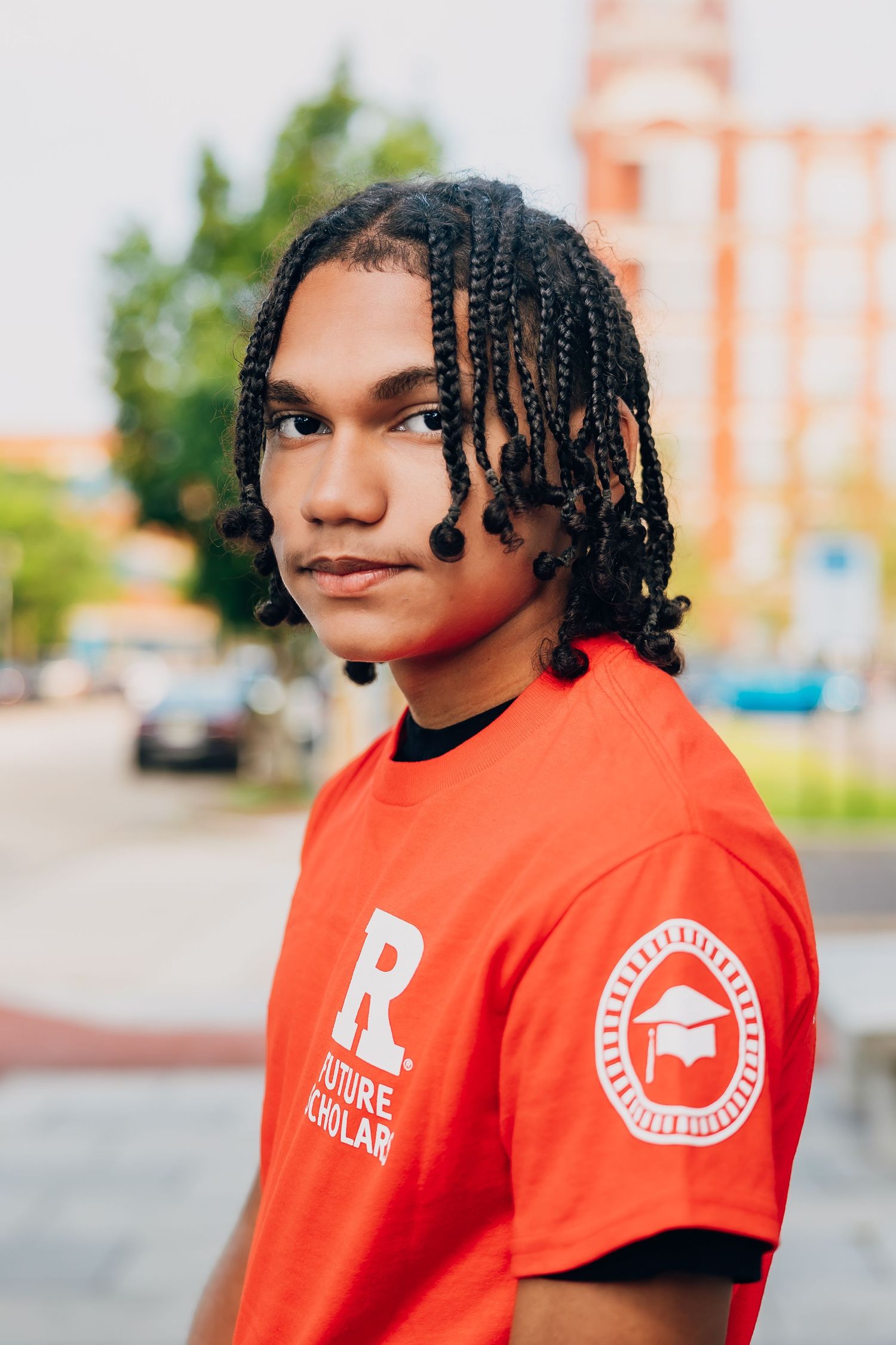 Young man with braids wearing a red Rutgers Future Scholars t-shirt outdoors on a urban street.