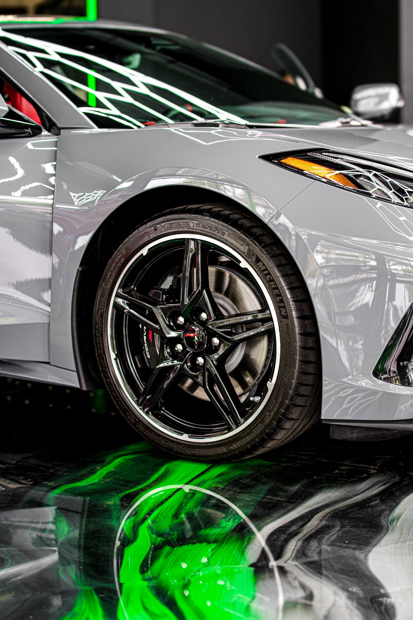 Silver Corvette C8 with black multi-spoke wheels on a reflective floor with green lighting accents.