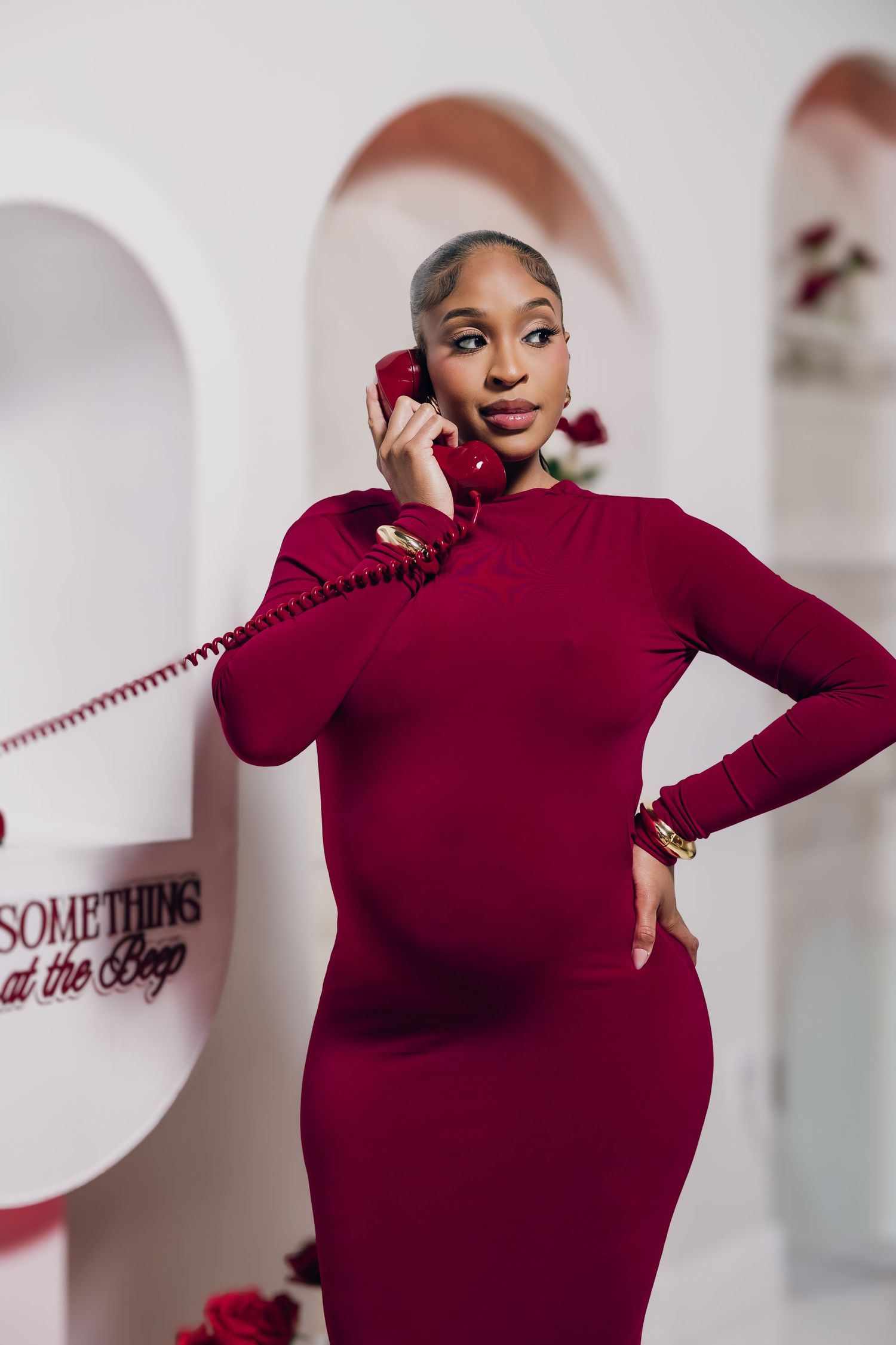 Pregnant woman in burgundy dress holding red vintage phone, posing elegantly in a white arched studio setting.