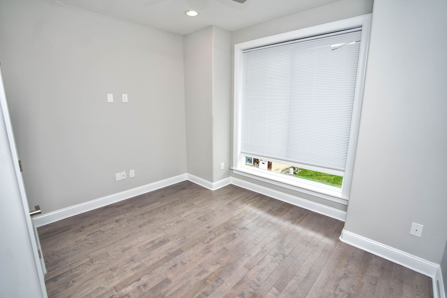 Empty room with light gray walls, wood-look flooring, and a large window with white blinds allowing natural light.