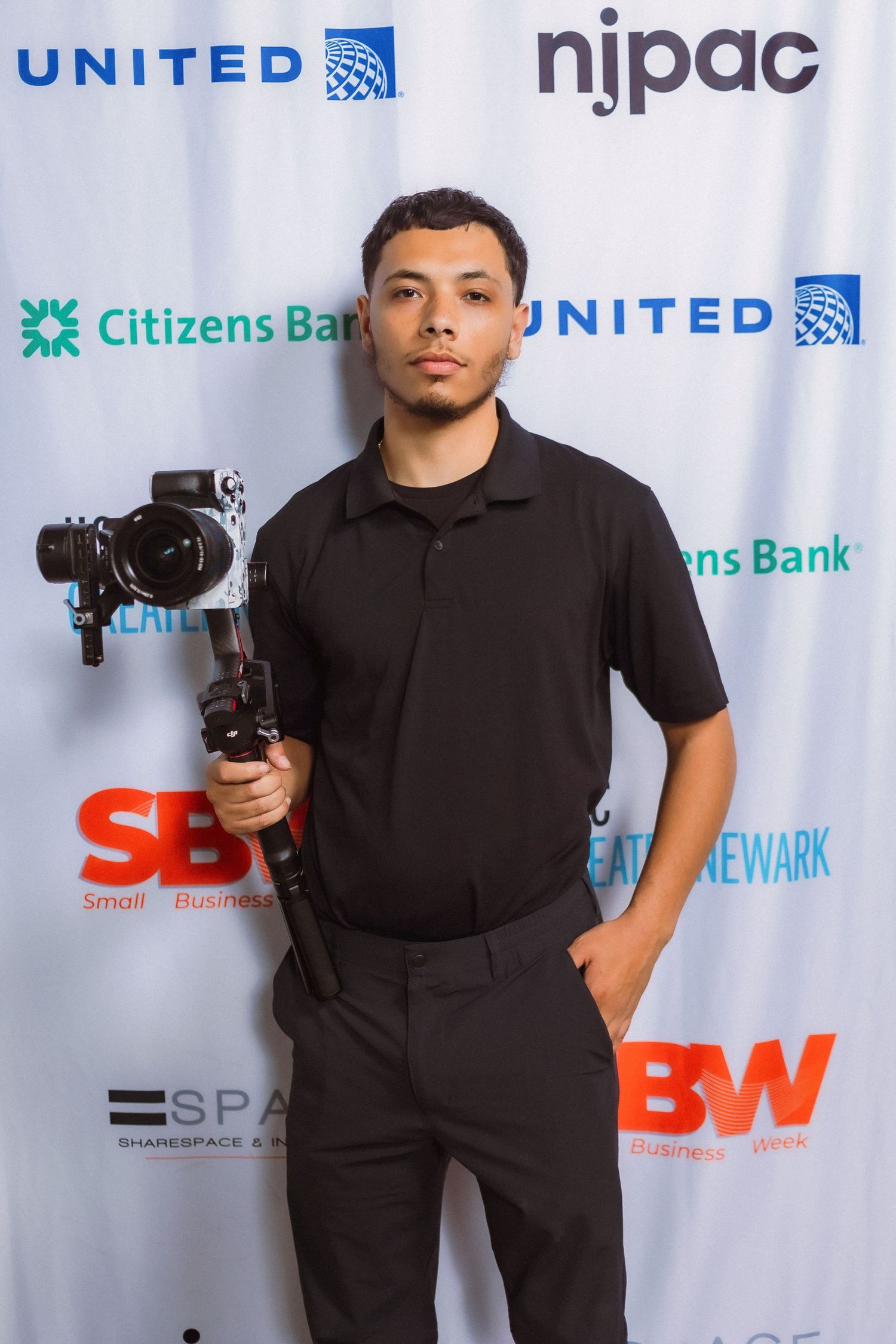 Professional videographer with camera rig standing against sponsor backdrop at corporate event.