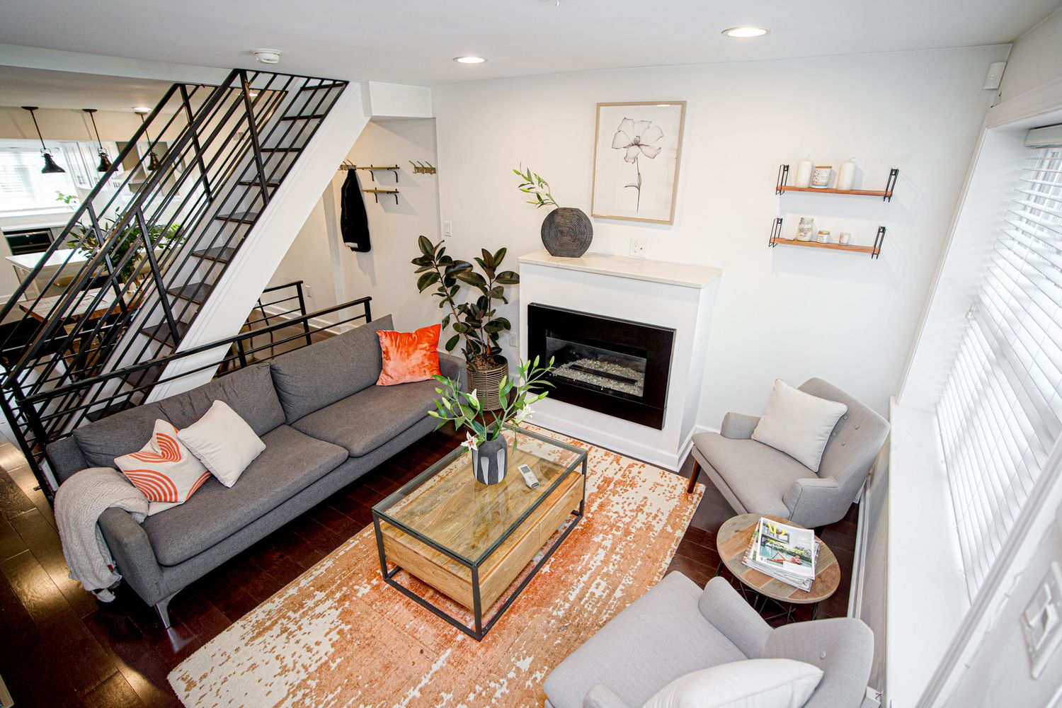 Modern living room with gray sofa, orange rug, glass coffee table, white fireplace, and industrial staircase.