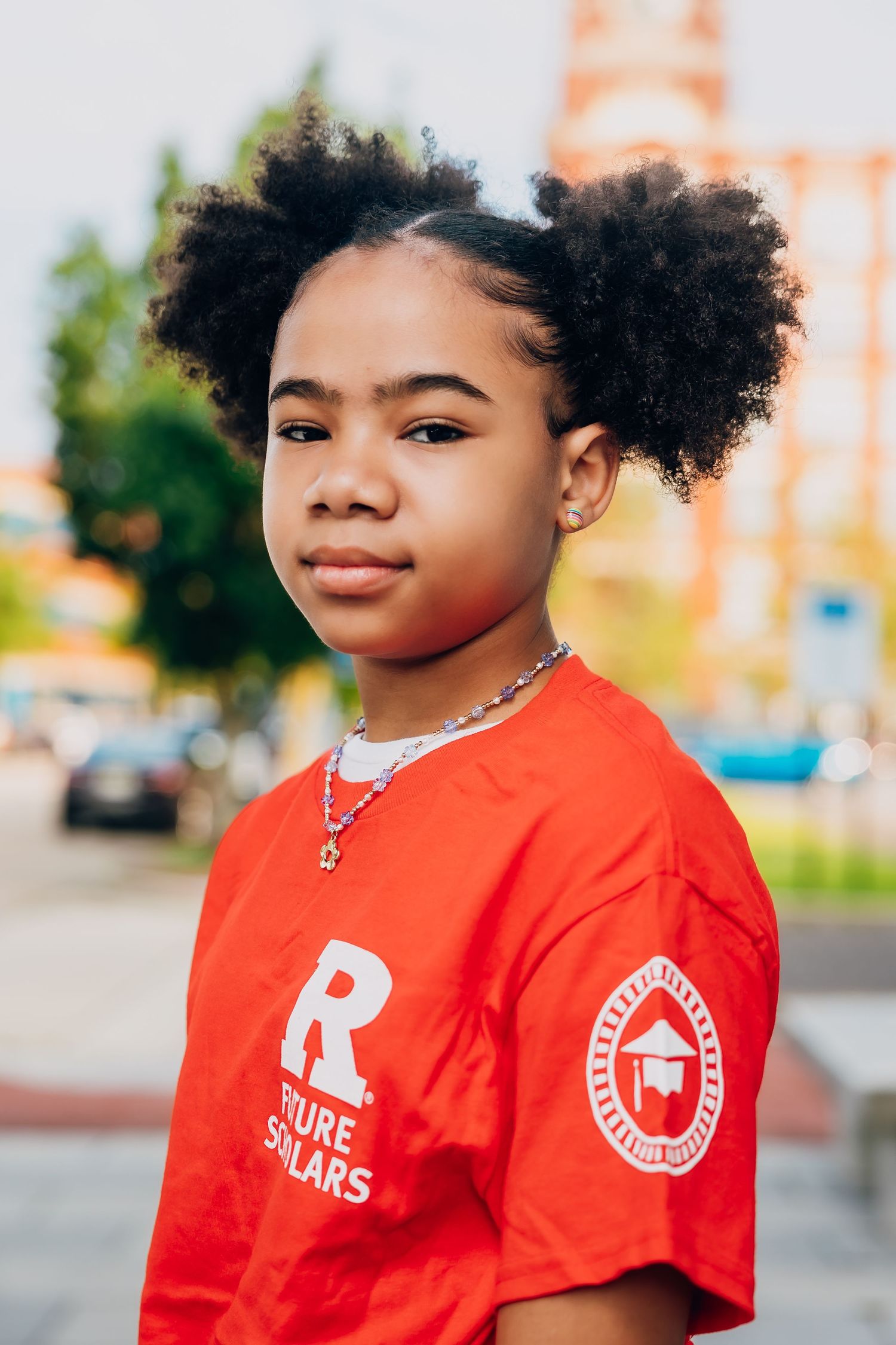 Young girl in red Future Solars t-shirt with afro puffs poses outdoors in urban setting.