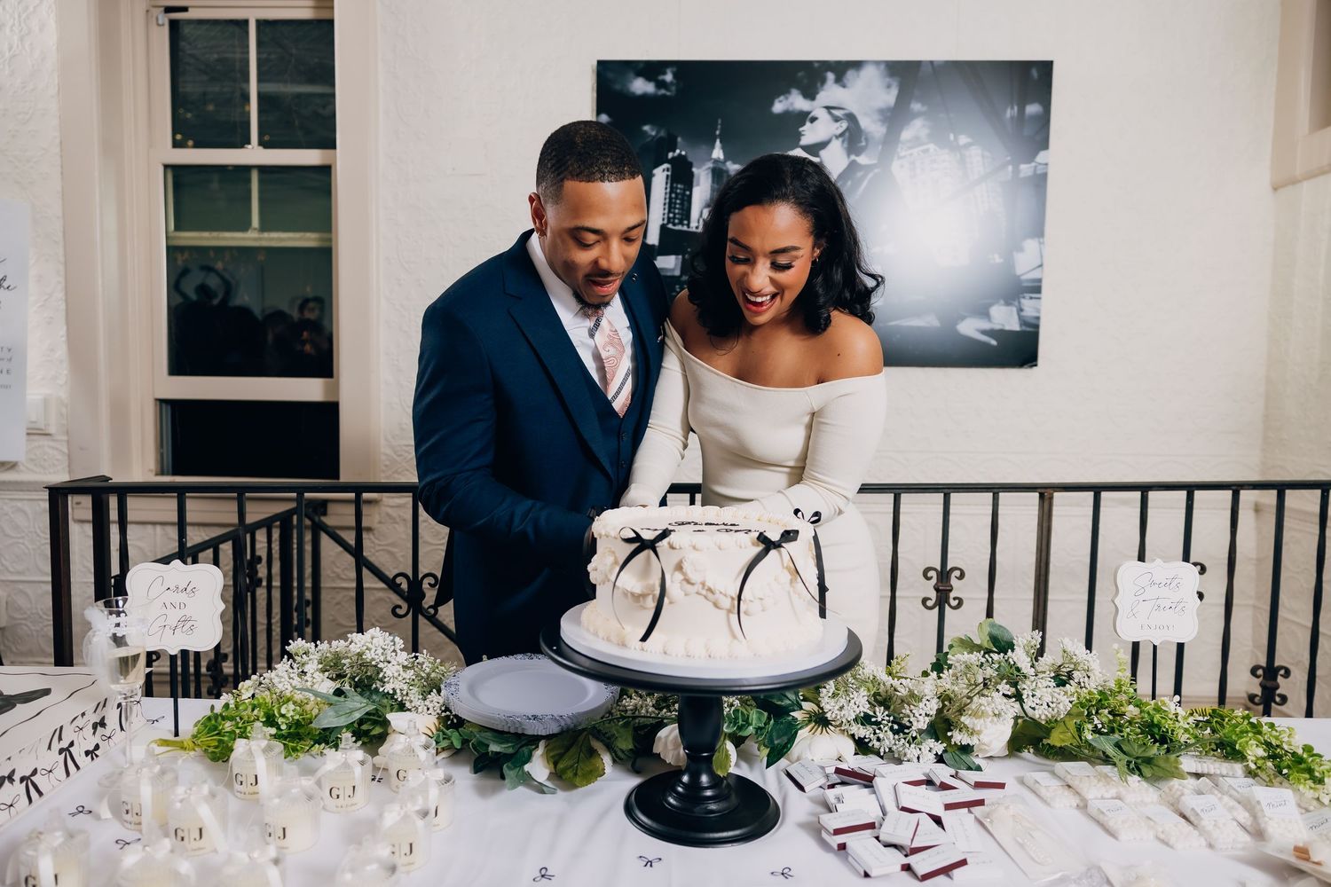 A couple cuts into their elegant black and white wedding cake surrounded by floral decorations at an indoor reception.