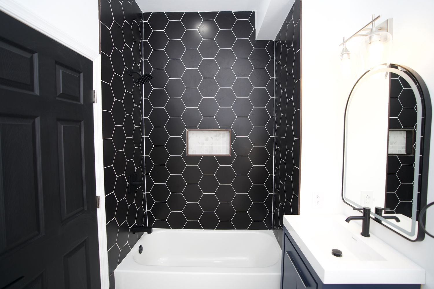 Modern bathroom featuring black hexagonal wall tiles, white bathtub, blue vanity, and arched mirror.