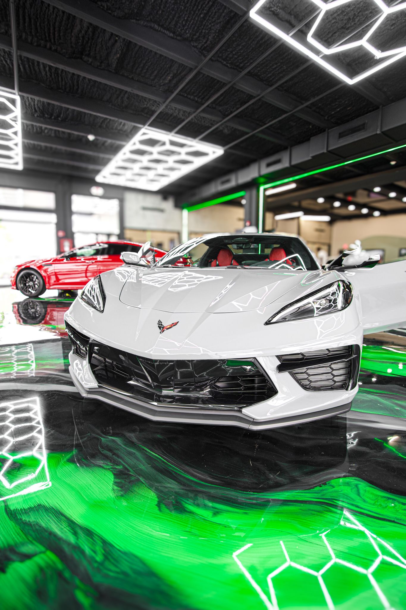 White Corvette C8 sports car displayed in a luxury showroom with green neon hexagonal lighting and reflective floors.