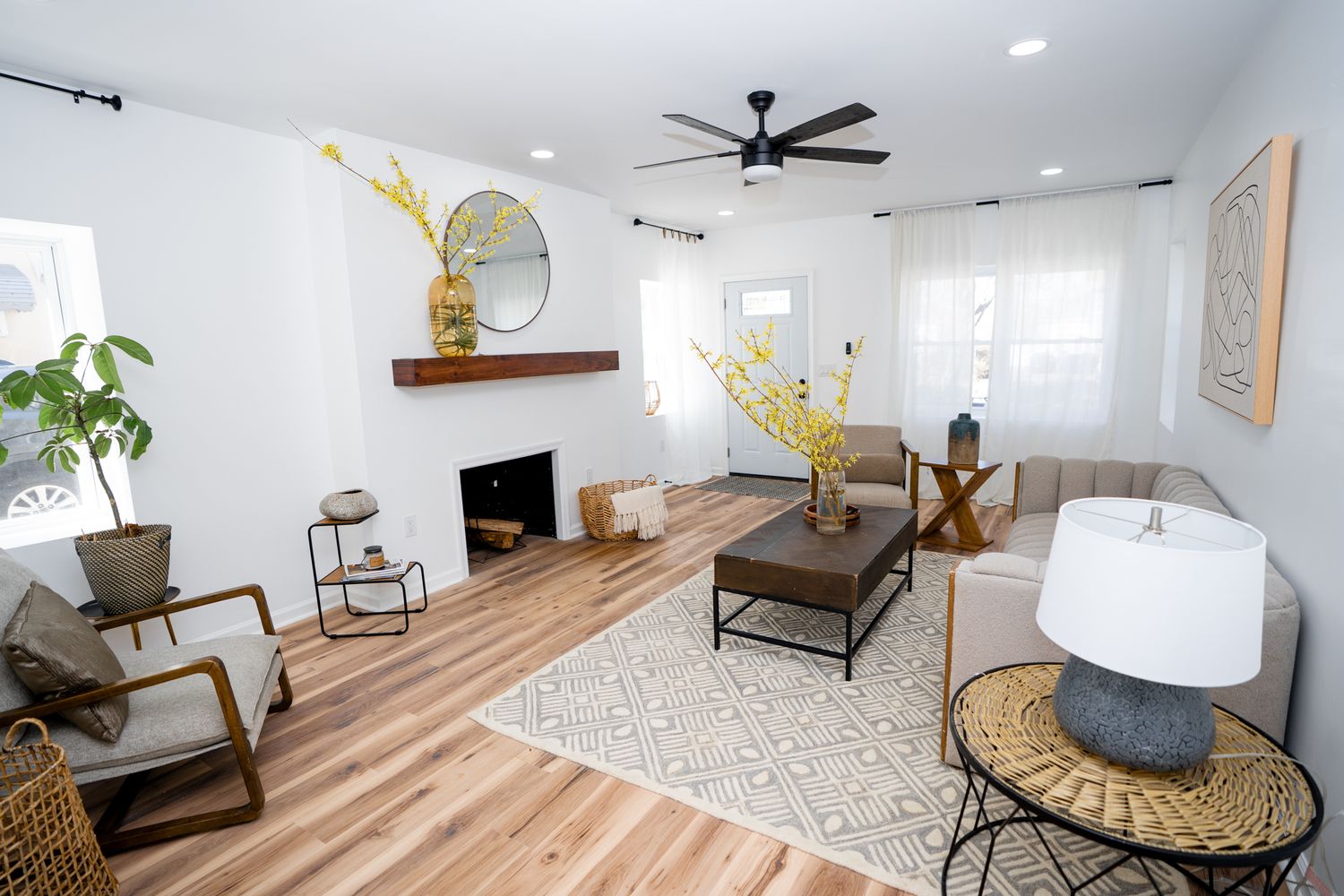 Bright living room with hardwood floors, white fireplace, ceiling fan, neutral sofa, and yellow floral accents.