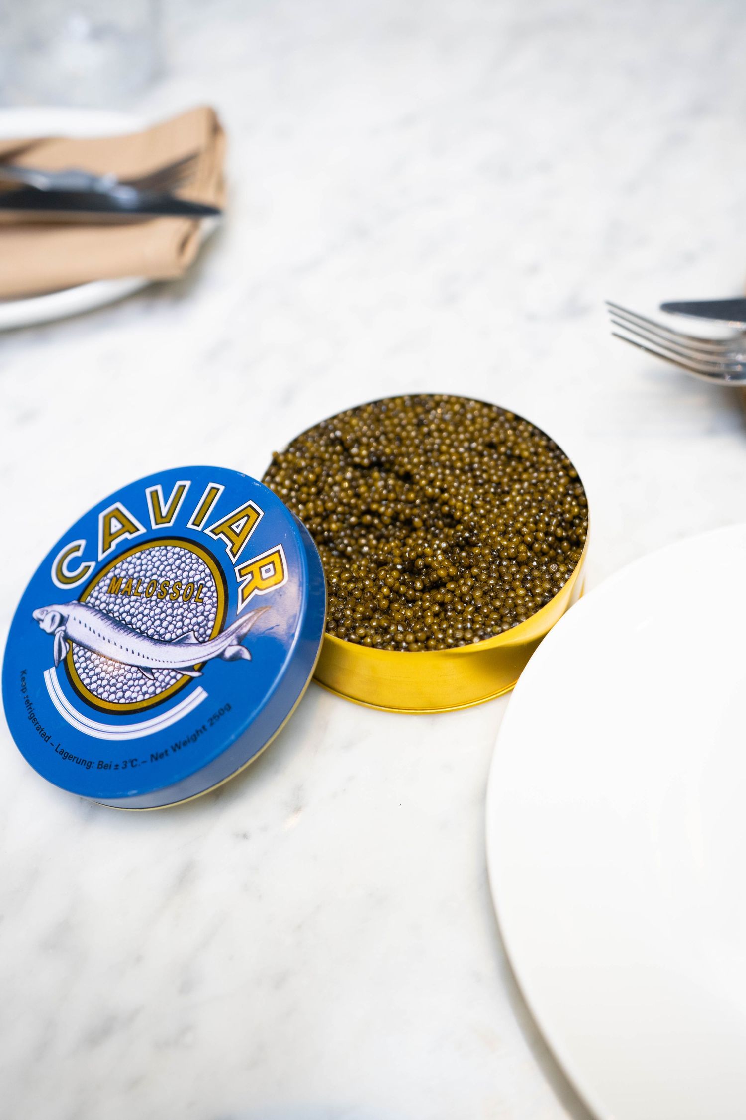 Open tin of black caviar on a marble surface, with a blue and gold label, beside a white plate.