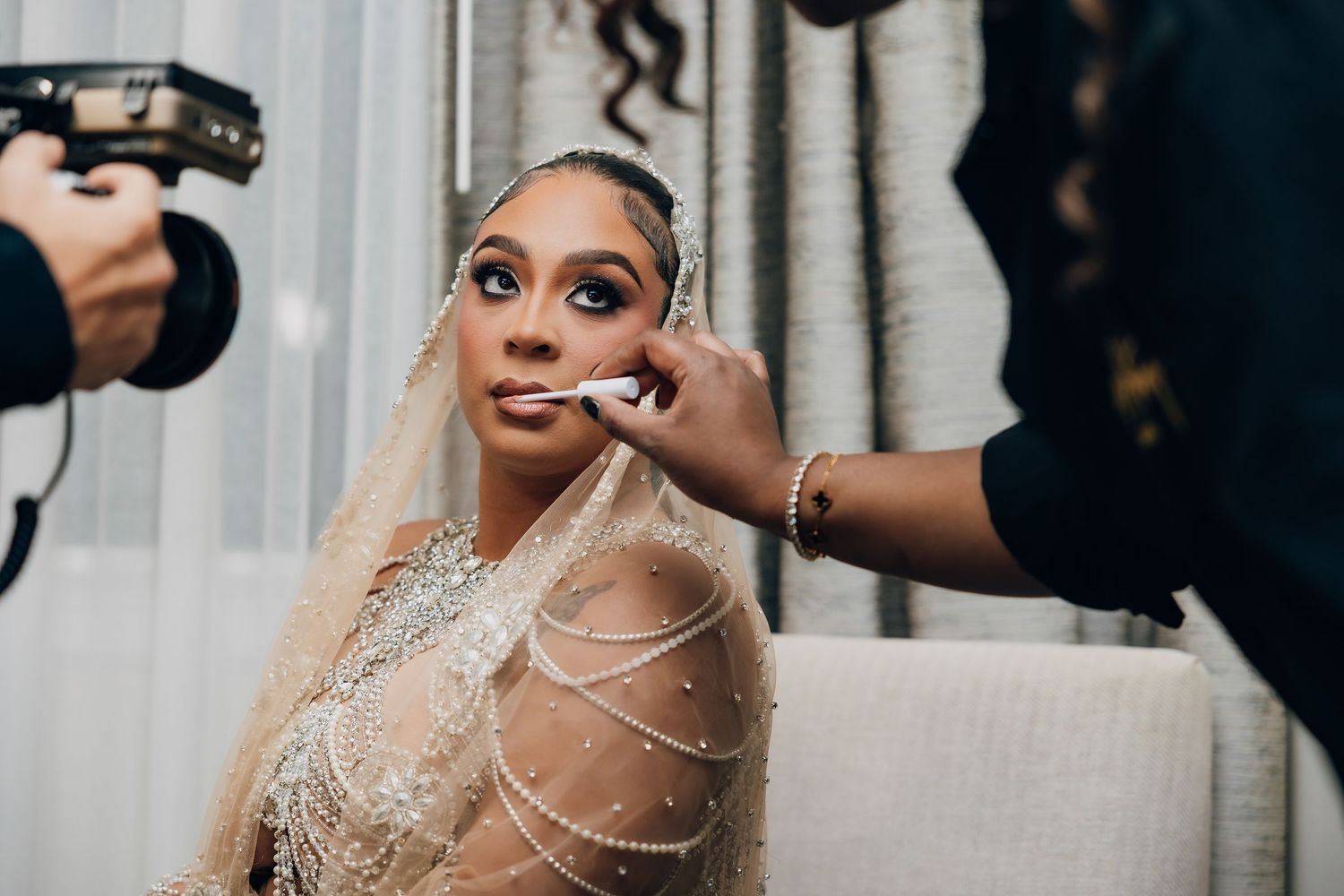 Bride getting her makeup professionally applied while wearing an embellished gown and pearl necklace.