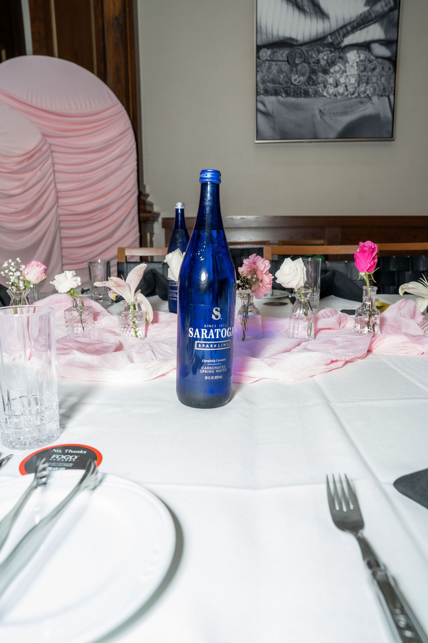 Blue Saratoga sparkling water bottle centerpiece on white tablecloth with pink runner, small floral arrangements, and place settings.