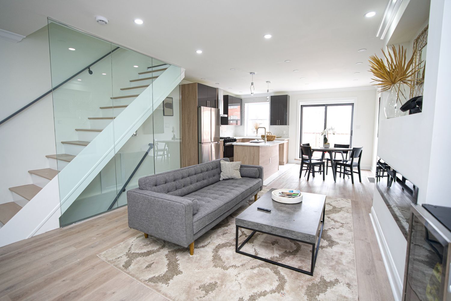 Modern open-concept living room with gray sofa, glass staircase, and kitchen in background on light hardwood floors.