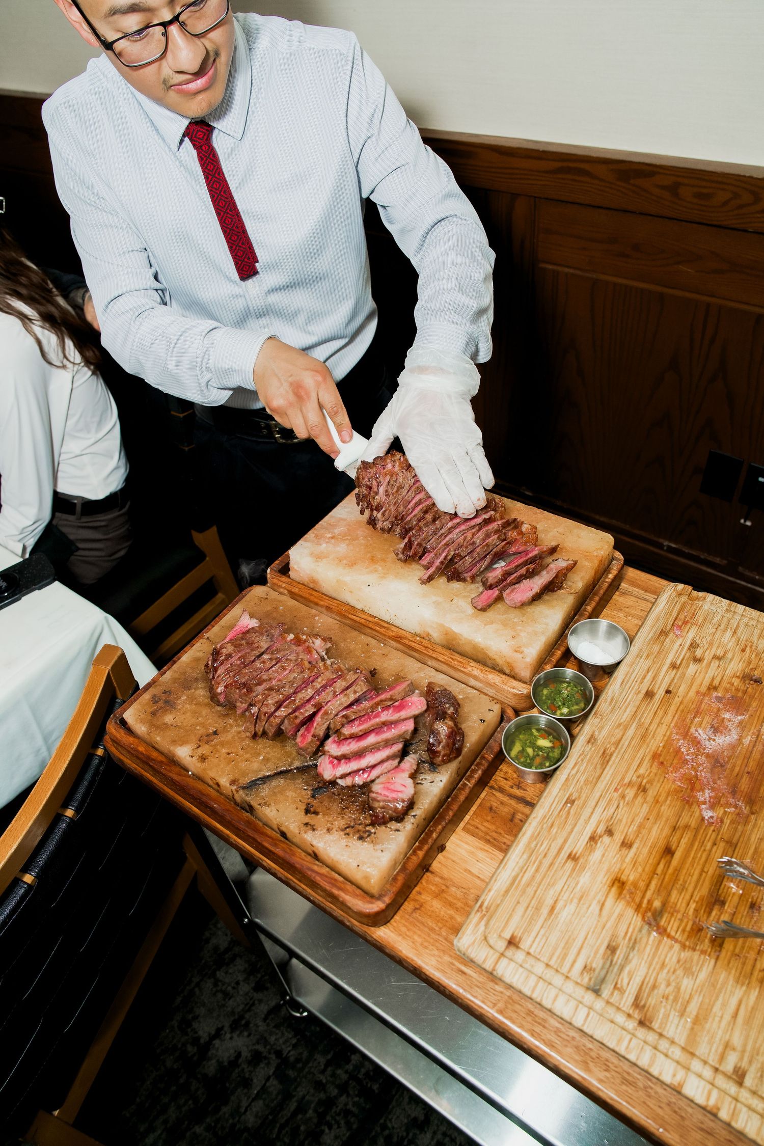 A waiter in white gloves slices medium-rare steak on wooden cutting boards with chimichurri sauce at a restaurant.