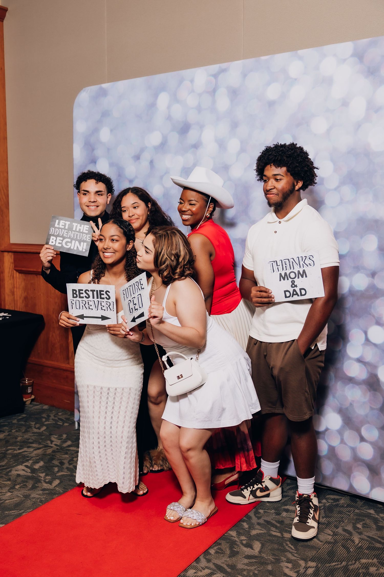 Group of six friends posing at a photo booth with fun signs at a graduation celebration on a red carpet.