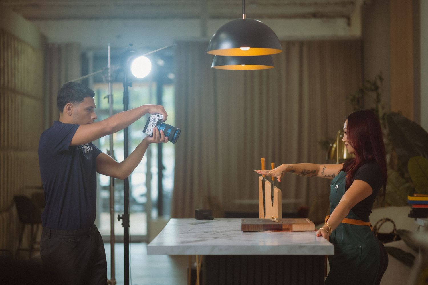 Two content creators filming a video in a modern kitchen setting with pendant lighting.