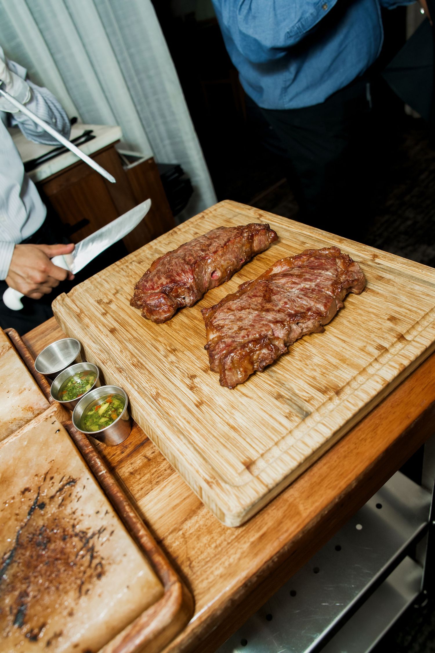 Chef slicing two grilled skirt steaks on a wooden cutting board with chimichurri sauce on the side.