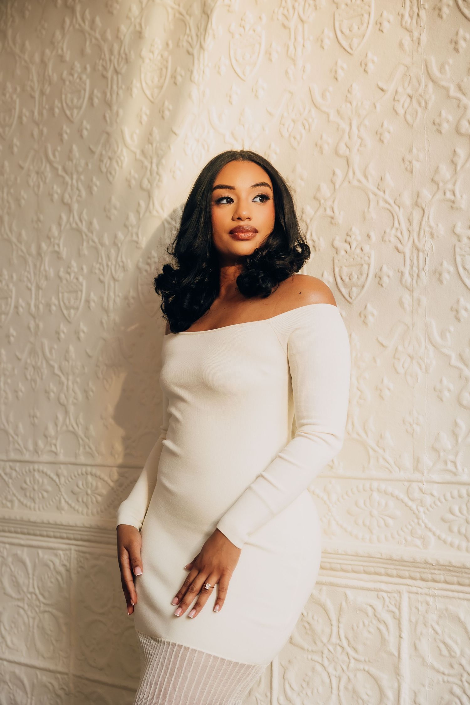 A young Black woman in a white off-shoulder bodycon dress poses against an ornate white textured wall.