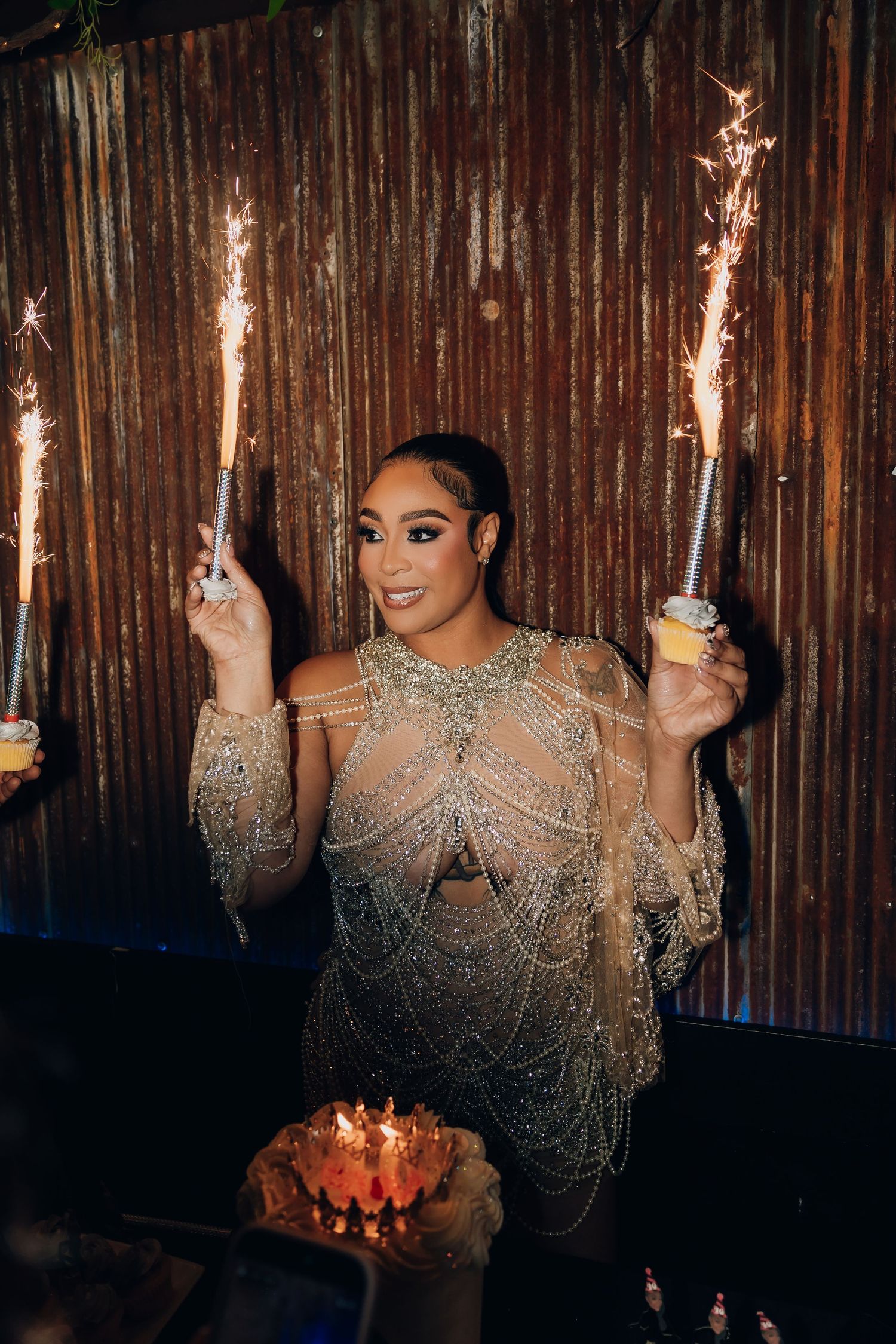 Woman in beaded gown holds sparklers beside birthday cake at glamorous celebration party.