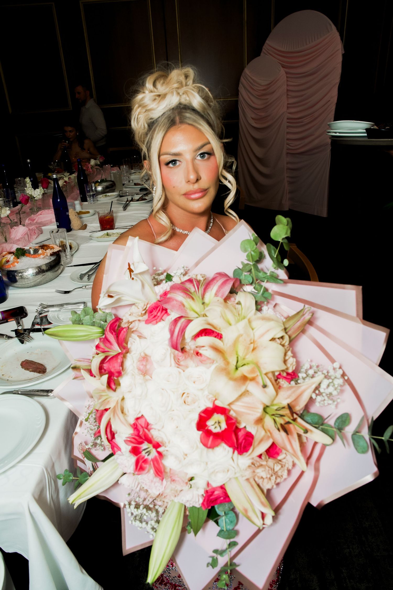 Elegant bride holding a large bouquet of pink lilies and white flowers at a wedding reception dinner table.