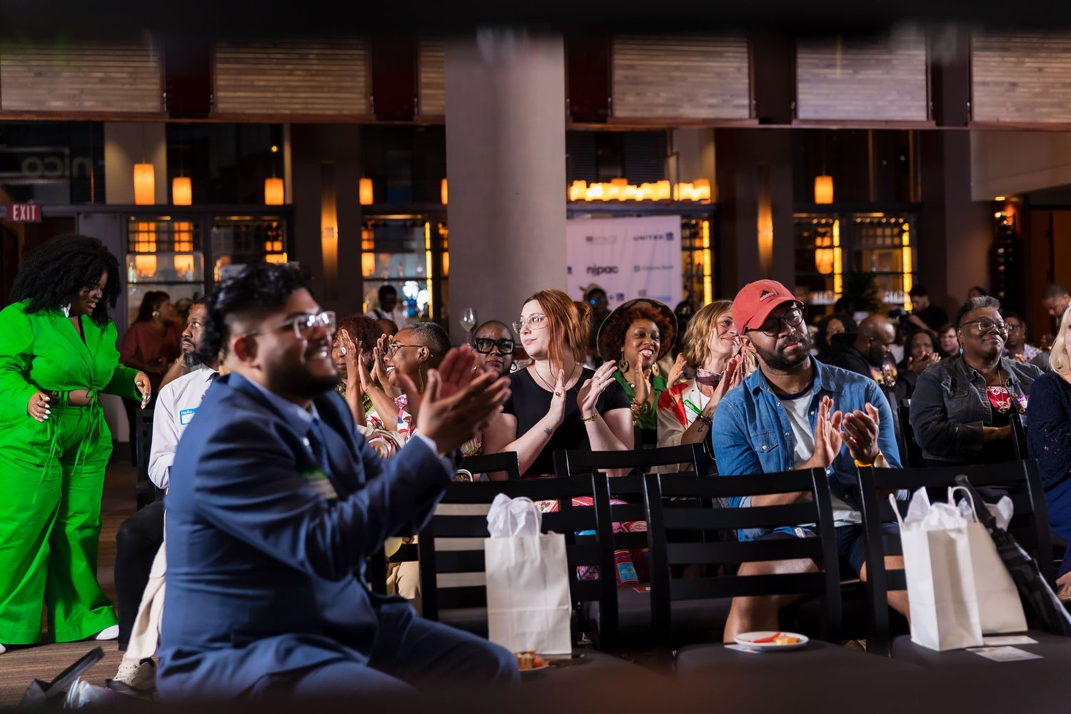 Audience members applauding enthusiastically during indoor event with shopping bags visible.
