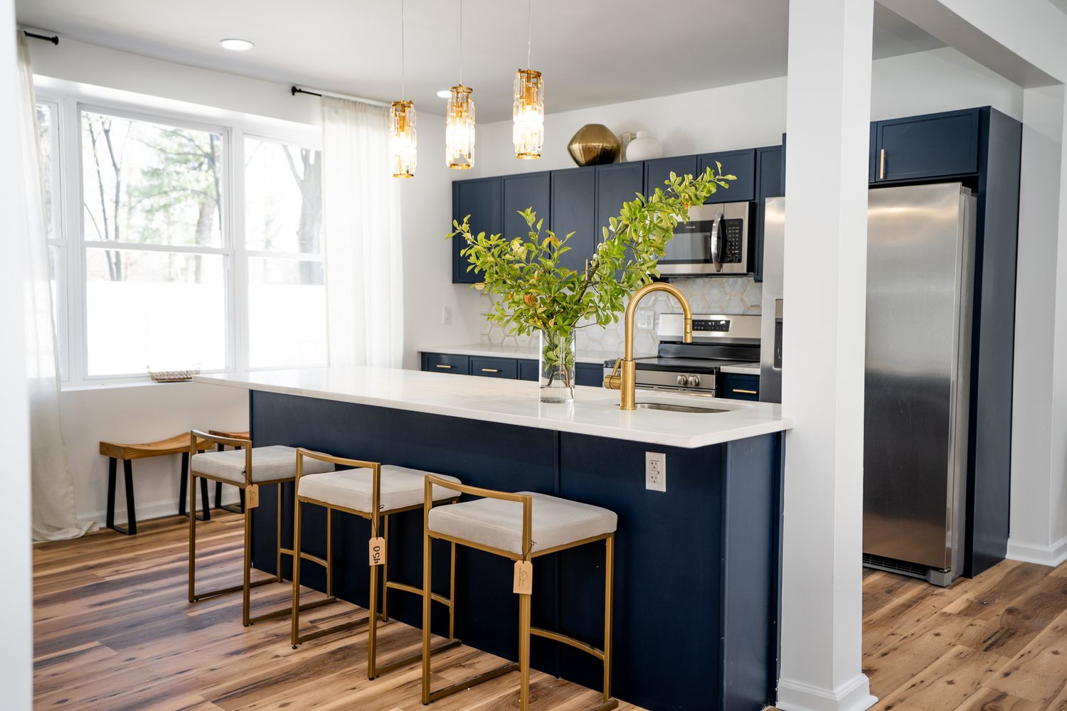 Modern navy blue kitchen with gold accents, white quartz island, bar stools, stainless appliances, and hardwood floors.