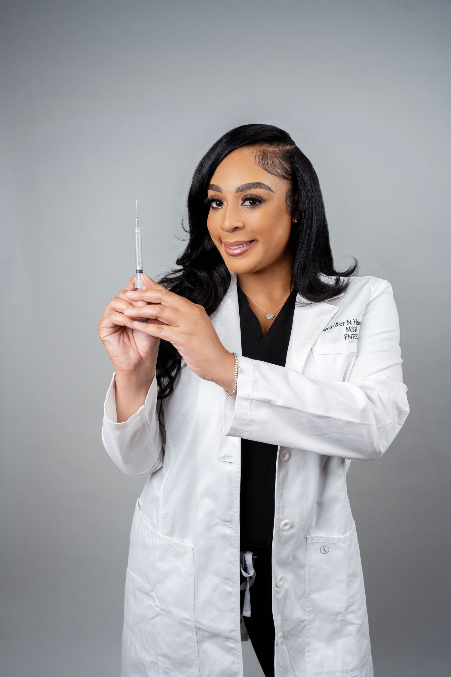 Female doctor in white lab coat holding a syringe, smiling confidently against a gray background.