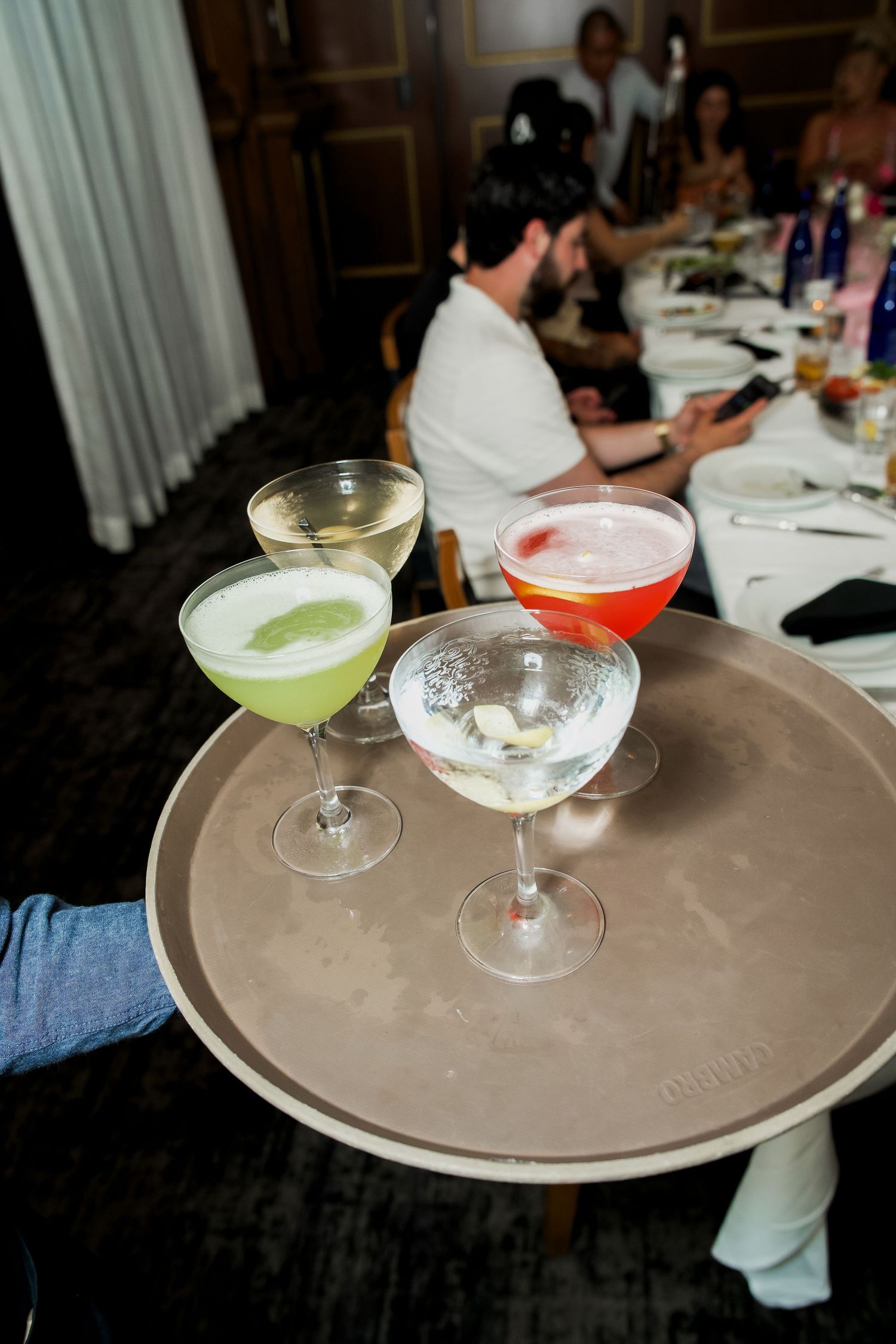 Server carrying a tray with three colorful cocktails including green, white, and pink drinks at an elegant dinner event.
