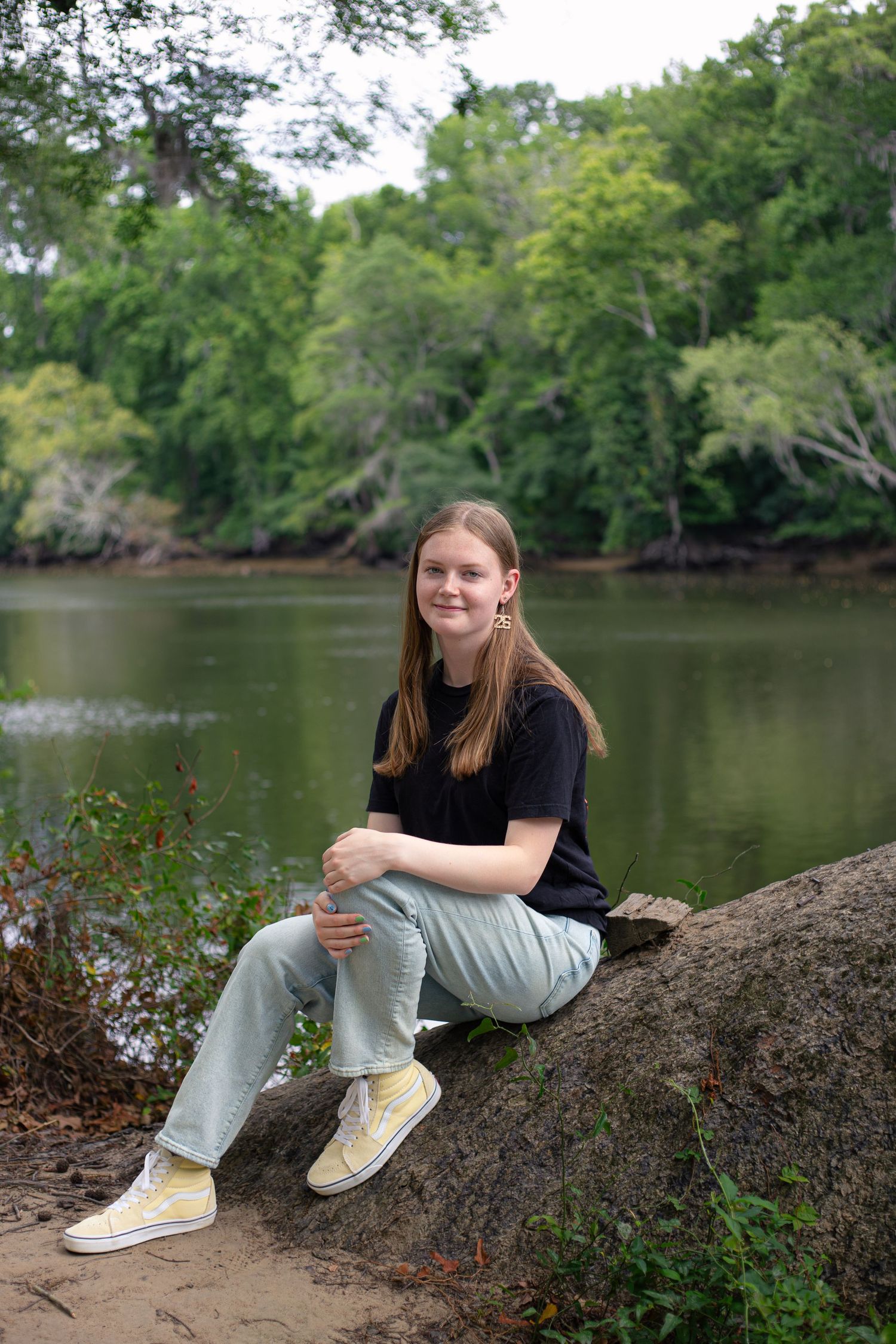 Young woman sitting on a rock by a calm river, wearing a black tee, light jeans, and yellow sneakers, surrounded by lush trees.