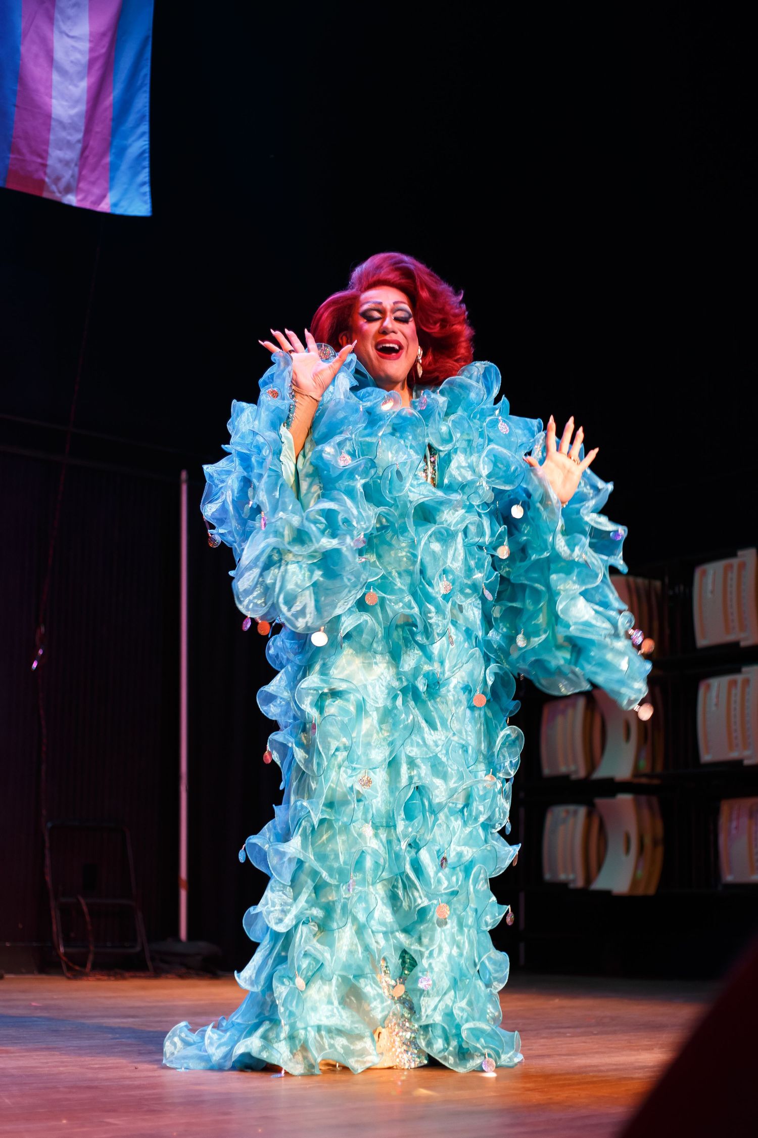 Performer in ruffled turquoise gown with red hair on stage, arms raised, with transgender pride flag visible.