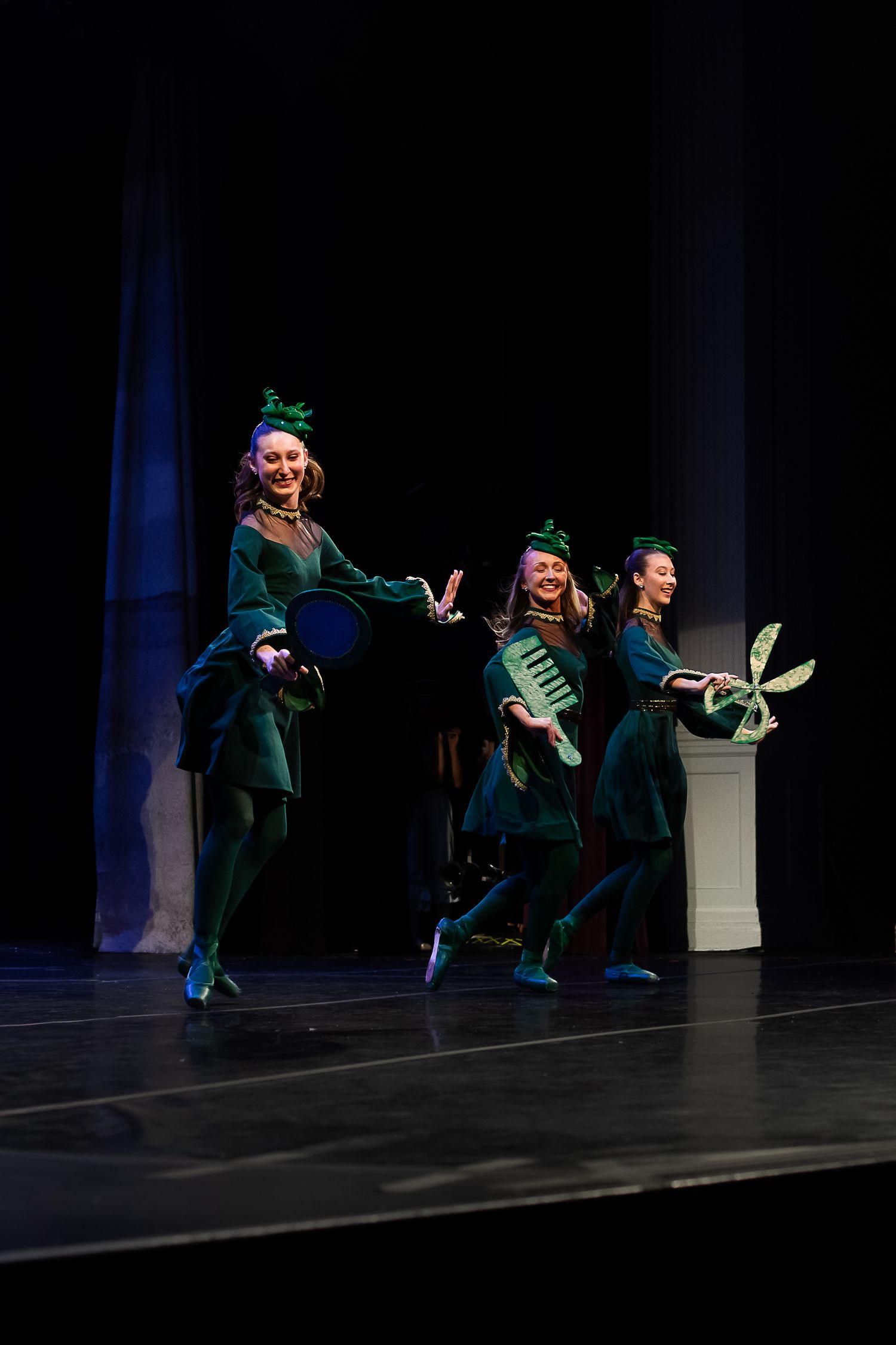 Three performers in green costumes and hats dance on stage during a theatrical production.