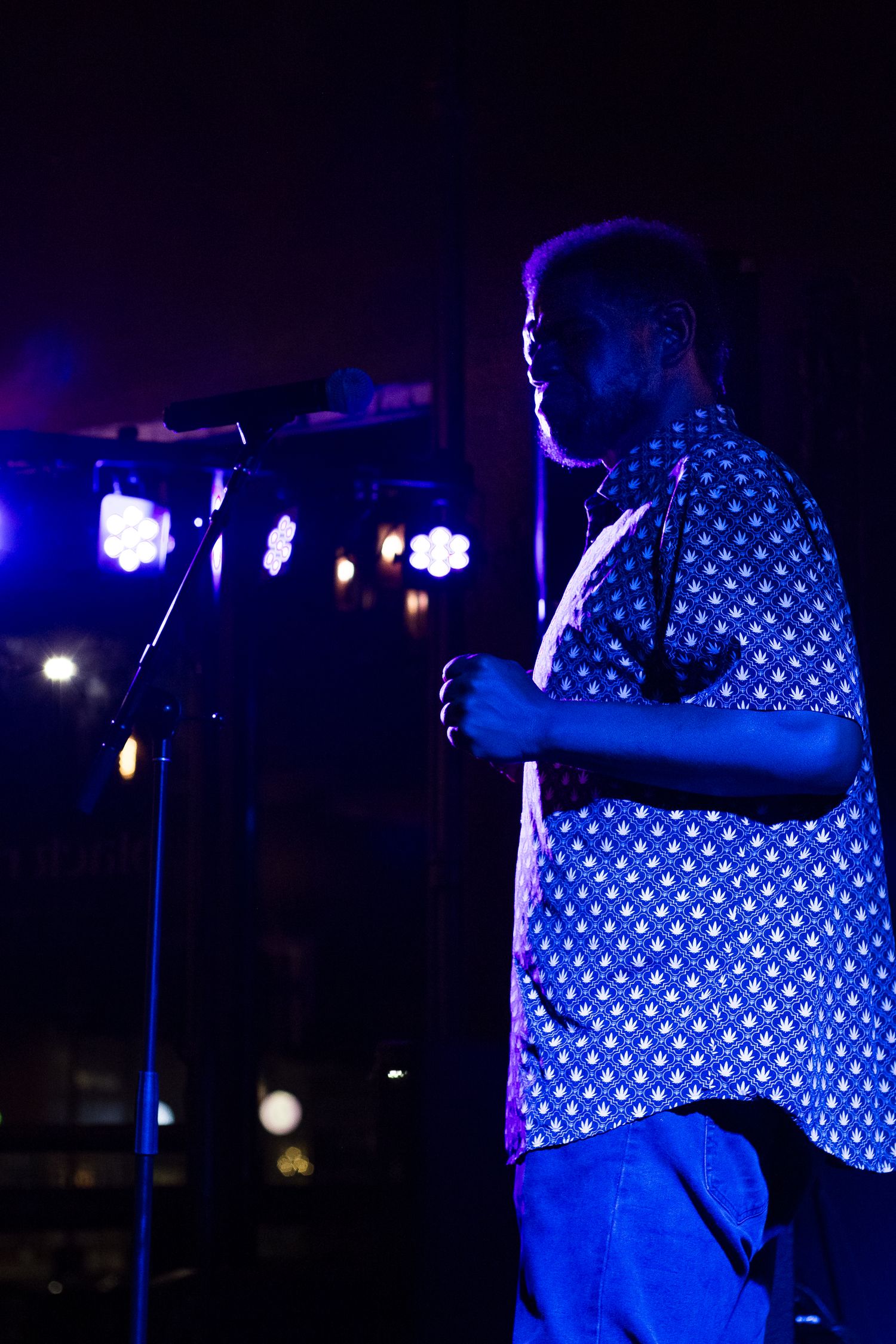 A man in a patterned blue shirt performs on stage at a dimly lit concert venue with colorful lights.