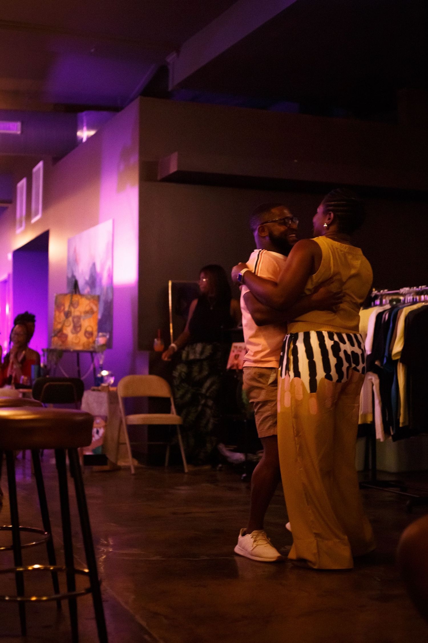 Two people slow dancing in a dimly lit venue with purple lighting, clothing rack visible in background.