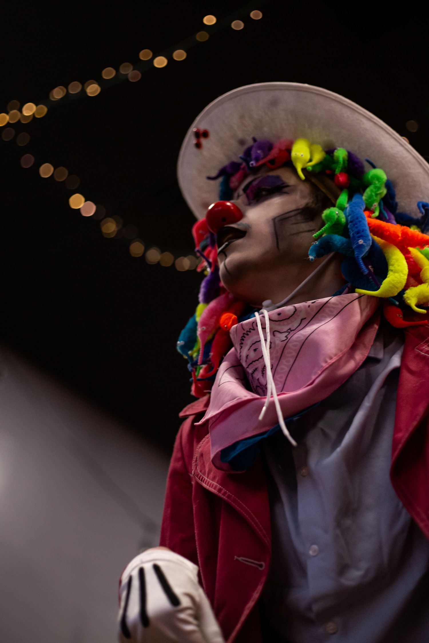 Clown costume with colorful wig, face paint, red jacket, and white hat under string lights.