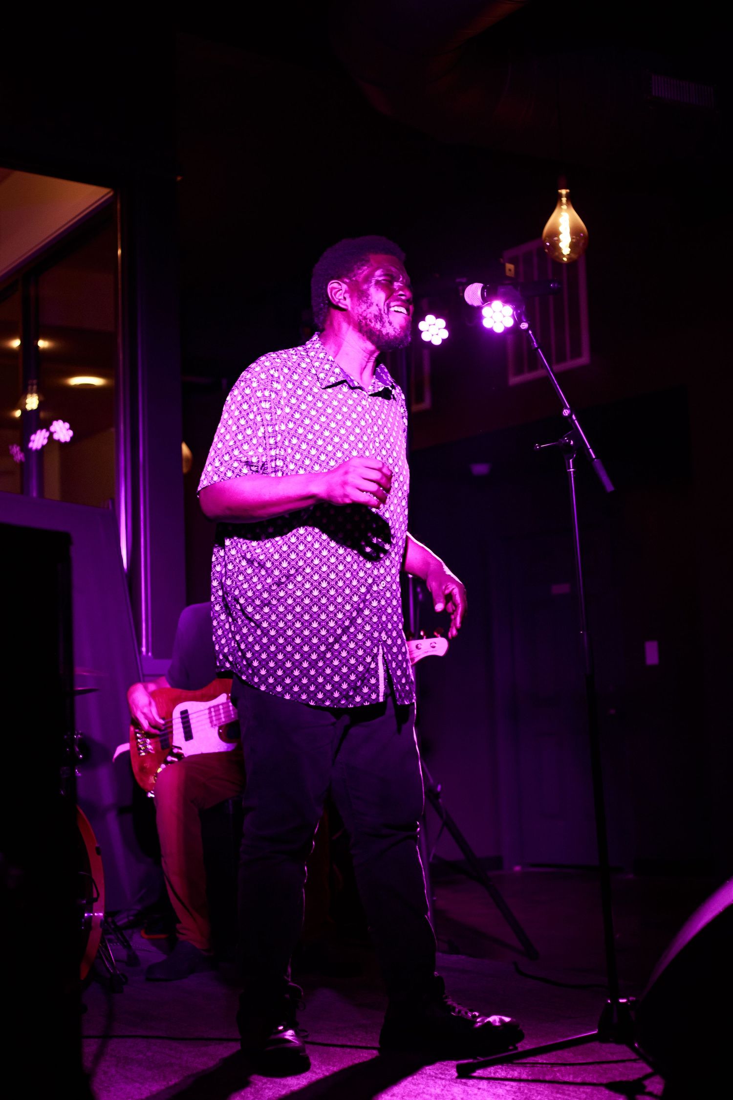 A man performs on stage under pink lights, wearing a patterned shirt, with a microphone stand and band equipment behind him.