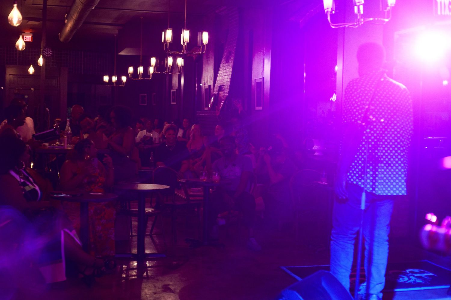 Comedian performing on stage under purple lights before a large crowd in a dimly lit venue.