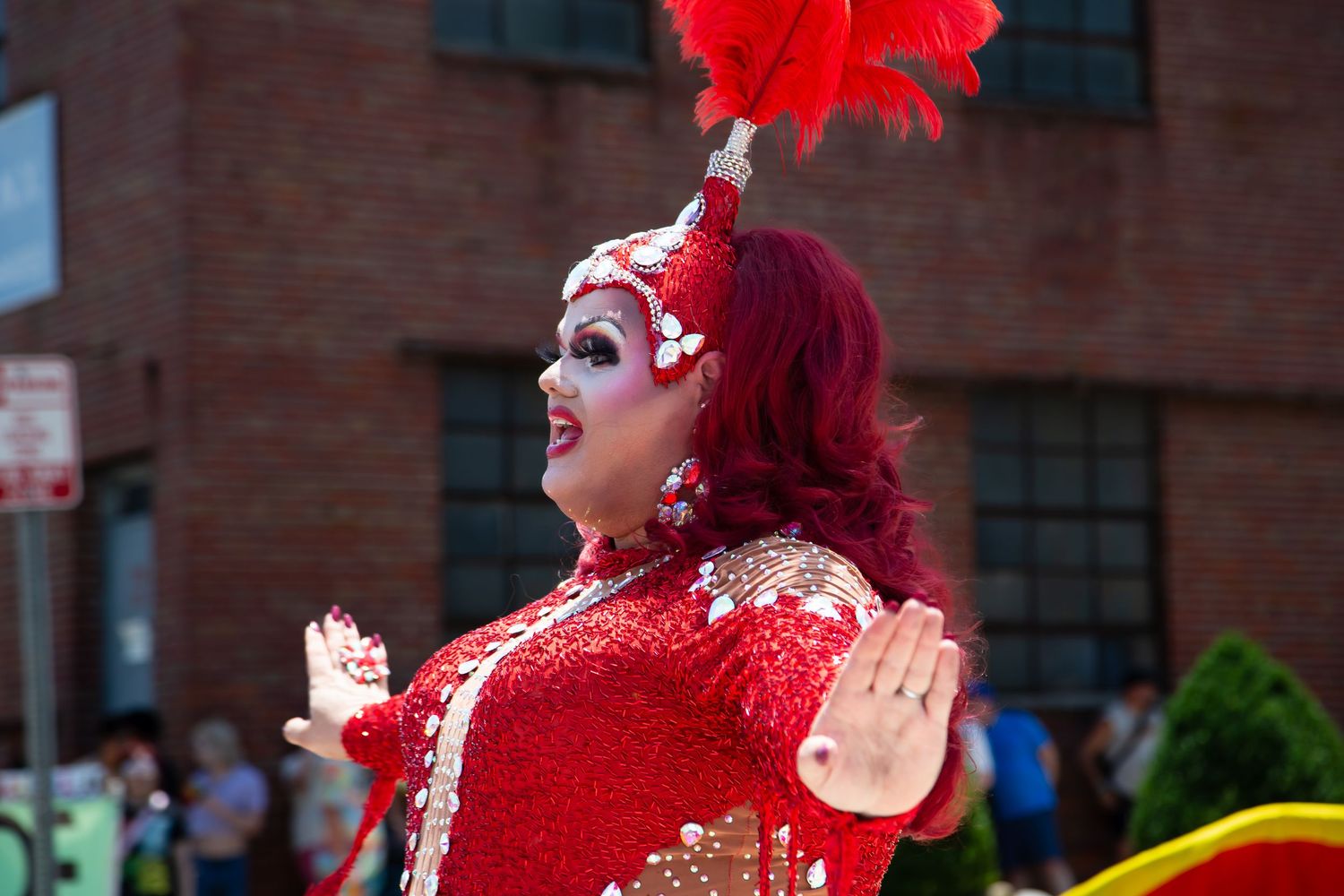 Drag performer in red sequin gown and feathered headpiece at a Pride parade.
