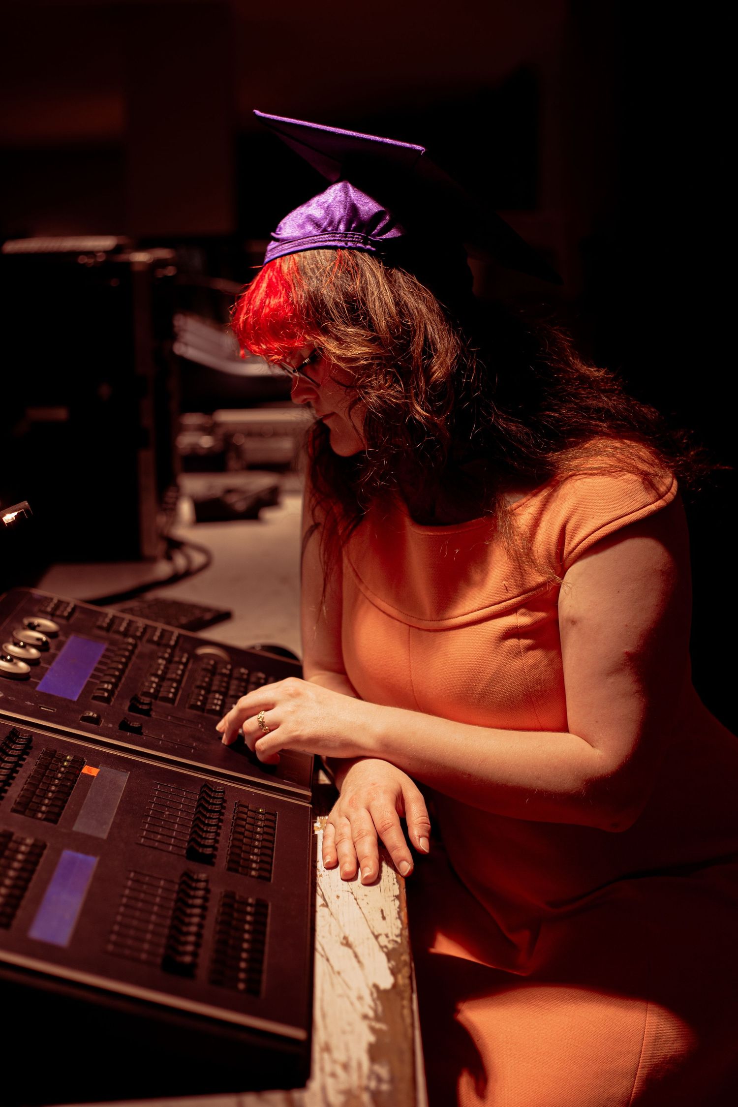 Woman in orange dress and graduation cap operating a professional audio mixing console in a dimly lit studio.