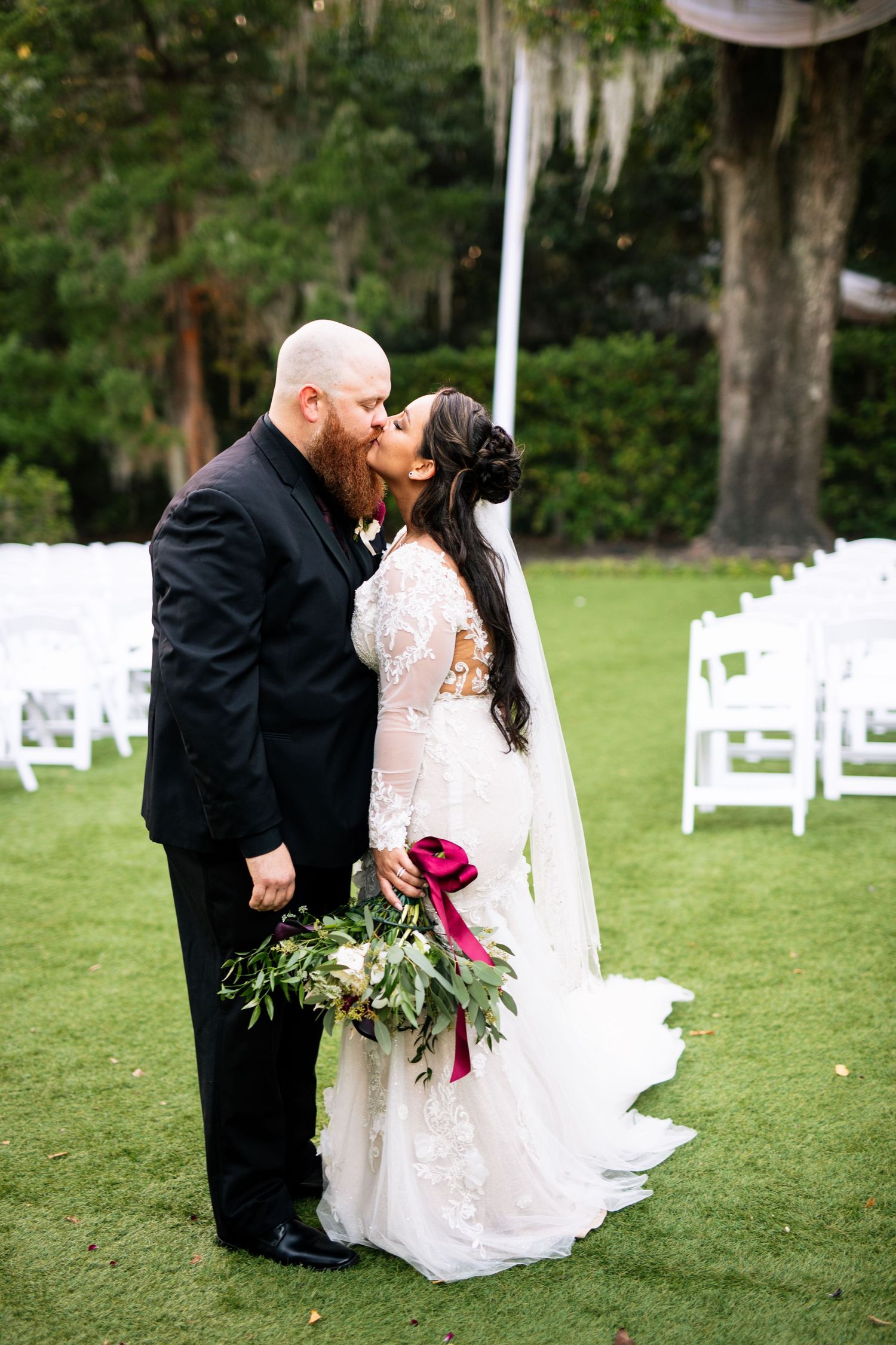 Bride and groom share a kiss on green lawn, white chairs behind them, bride holding lush bouquet with red flowers.