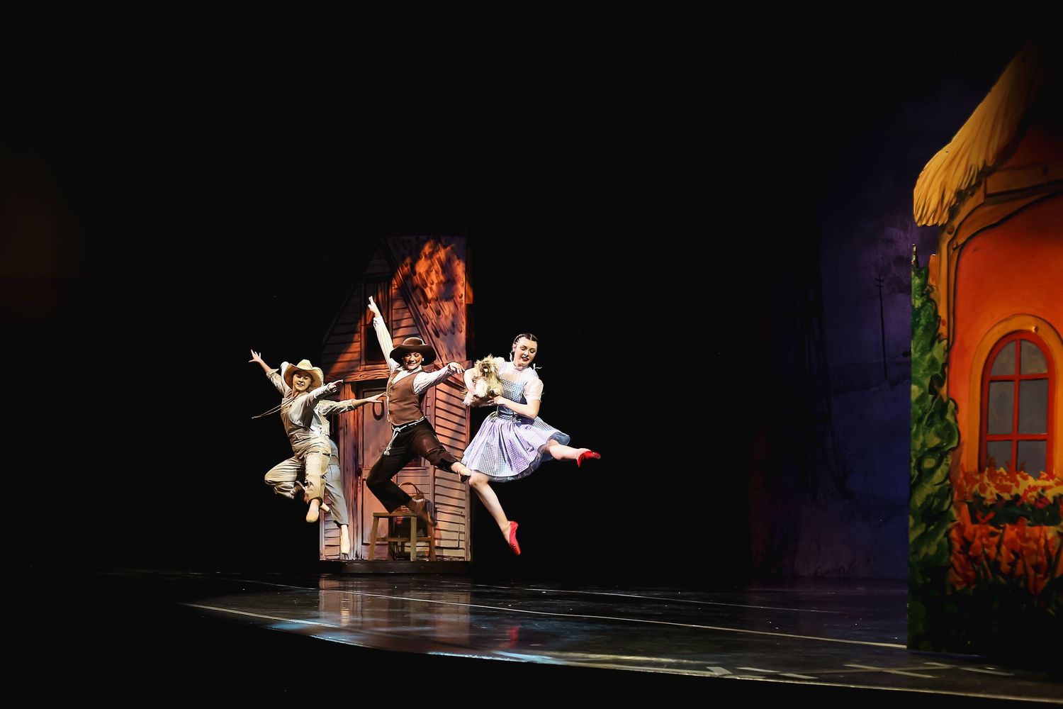 Performers leap across a theatrical stage in a colorful Wizard of Oz production with vibrant set design.