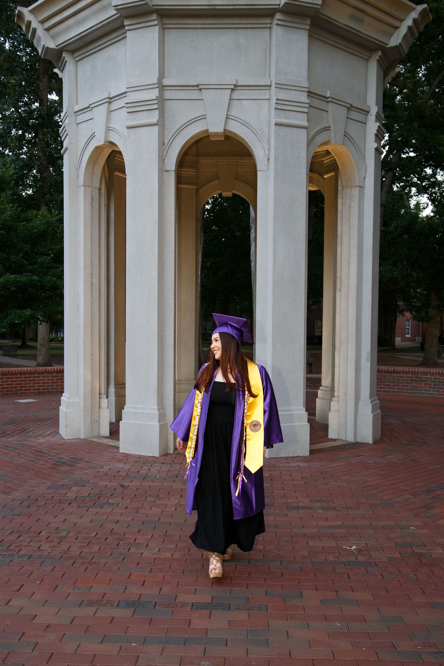 A young woman in purple graduation cap and gown with gold stoles walks in front of a white stone rotunda on brick pavement.