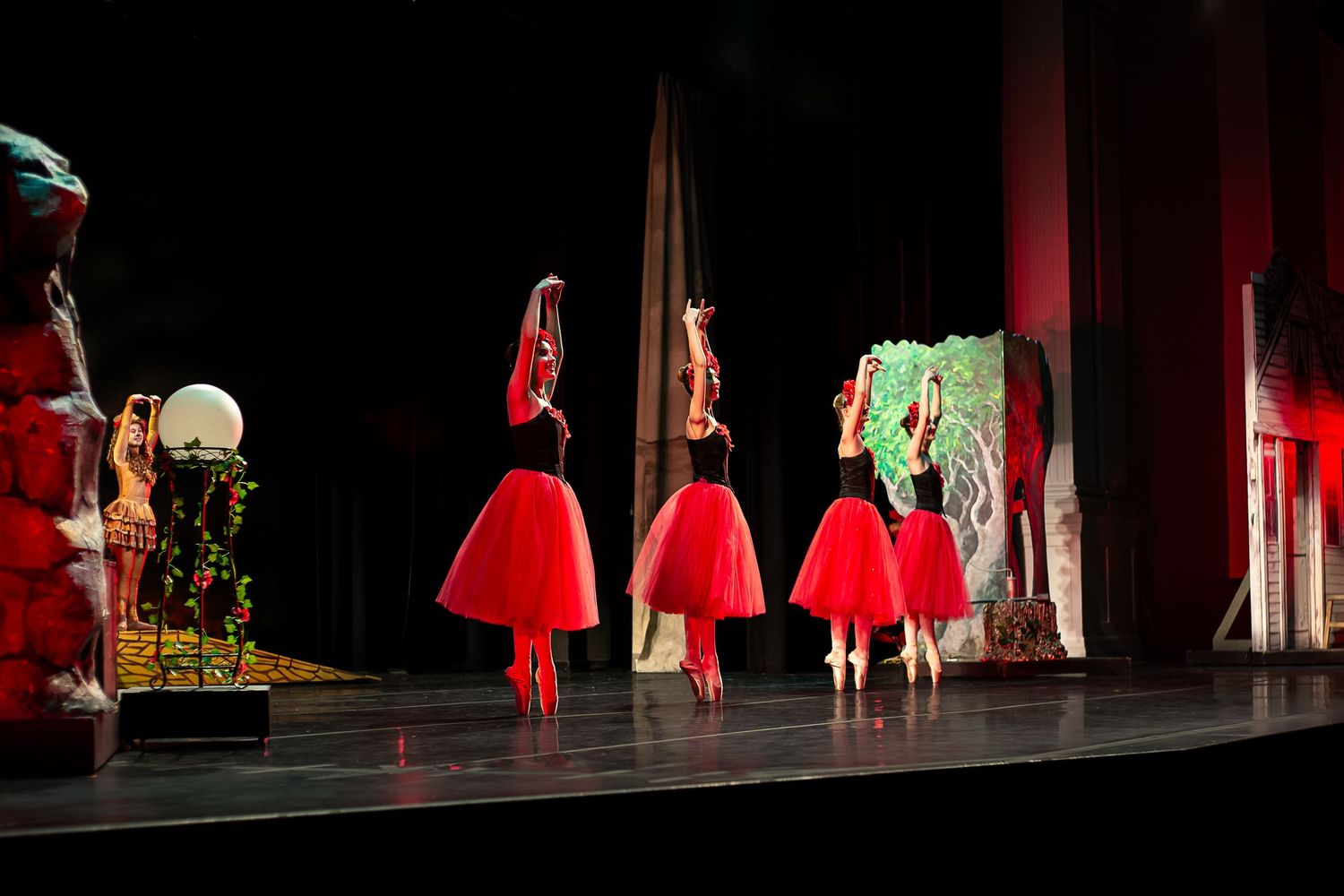 Ballet dancers in red tulle skirts perform gracefully on a dimly lit theater stage during a live dance recital.