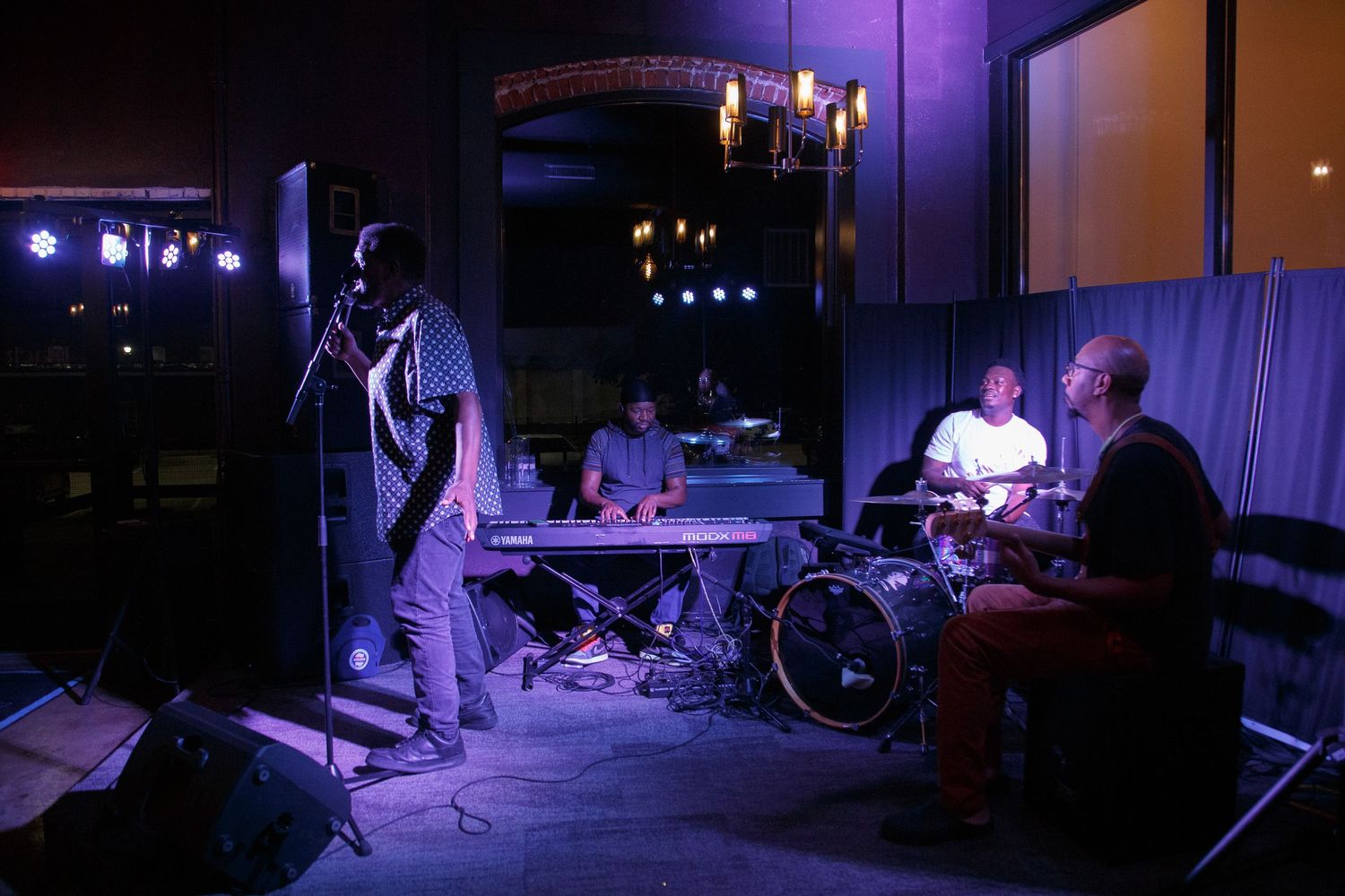 Live band performing on a dimly lit stage with purple lighting, featuring a vocalist, keyboardist, drummer, and guitarist.